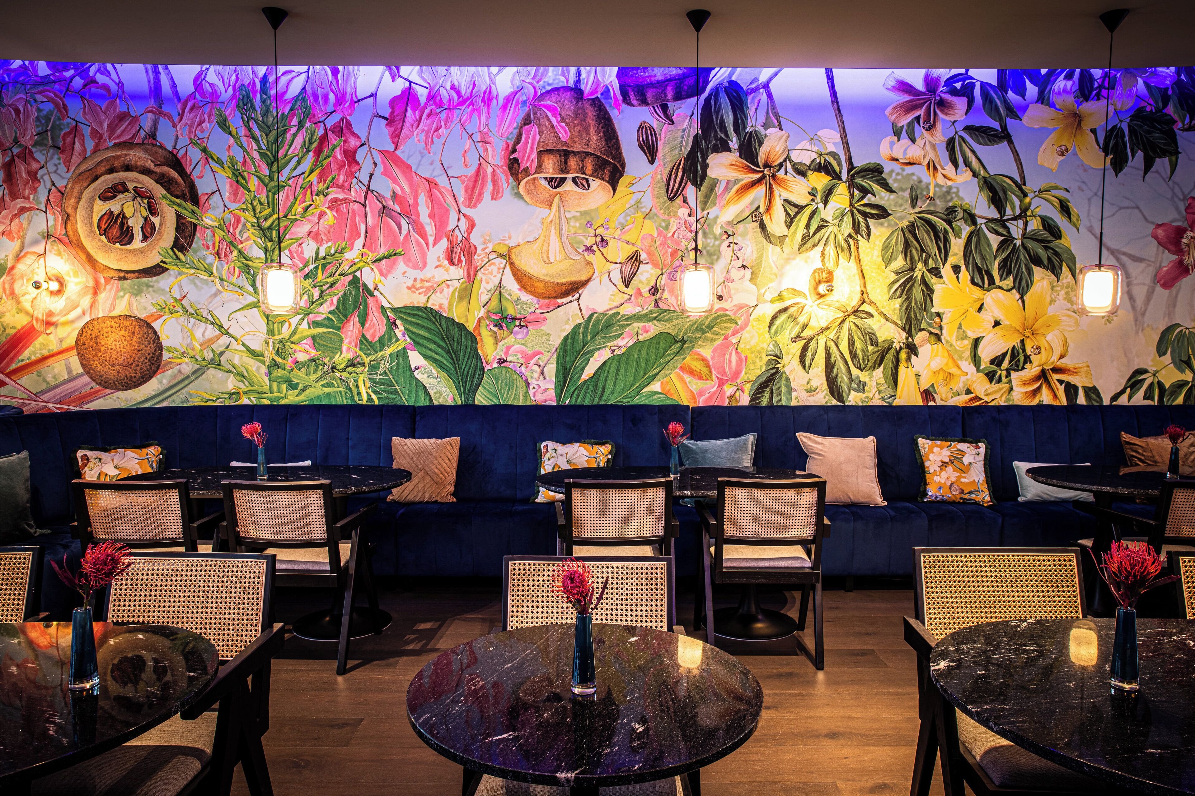Un restaurant agrémenté d’une fresque murale colorée, de tables rondes, de chaises en osier et d’un coin salon avec des sièges bleus et moelleux.