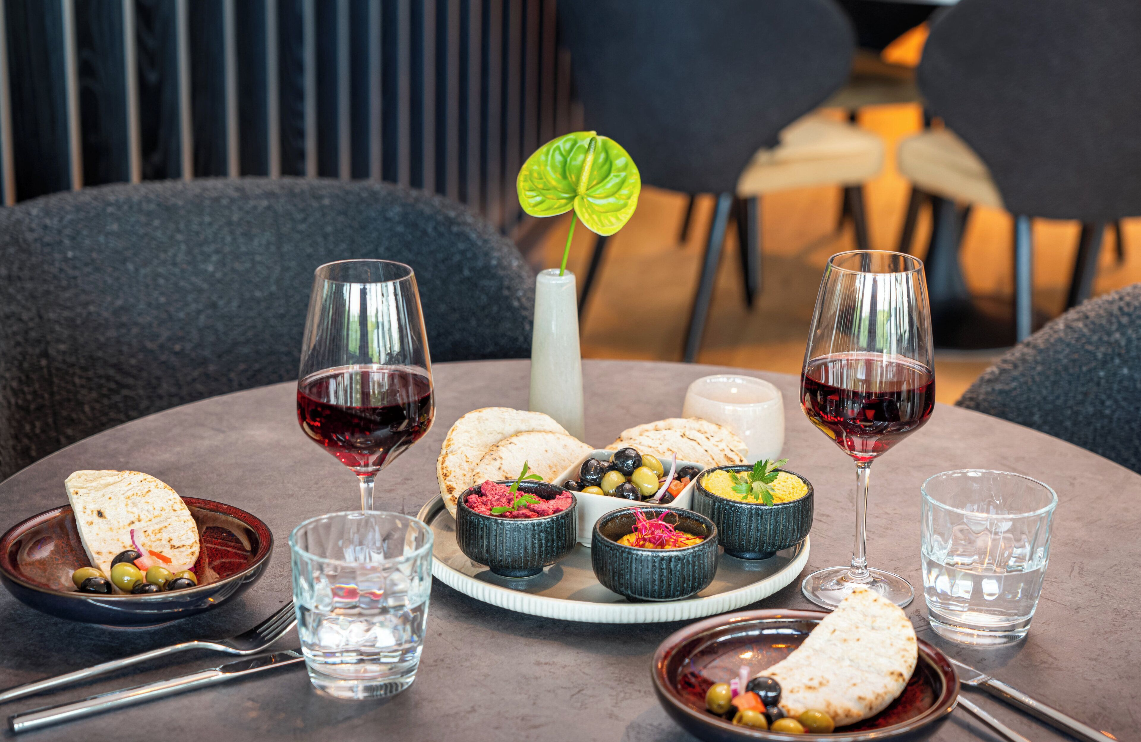 Una mesa de comedor con una variedad de platos, como pan plano, aceitunas y salsas, acompañados de copas de vino tinto.