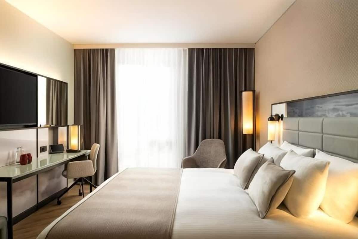 A hotel room with a bed, a desk, a chair, a TV, and a window with curtains.