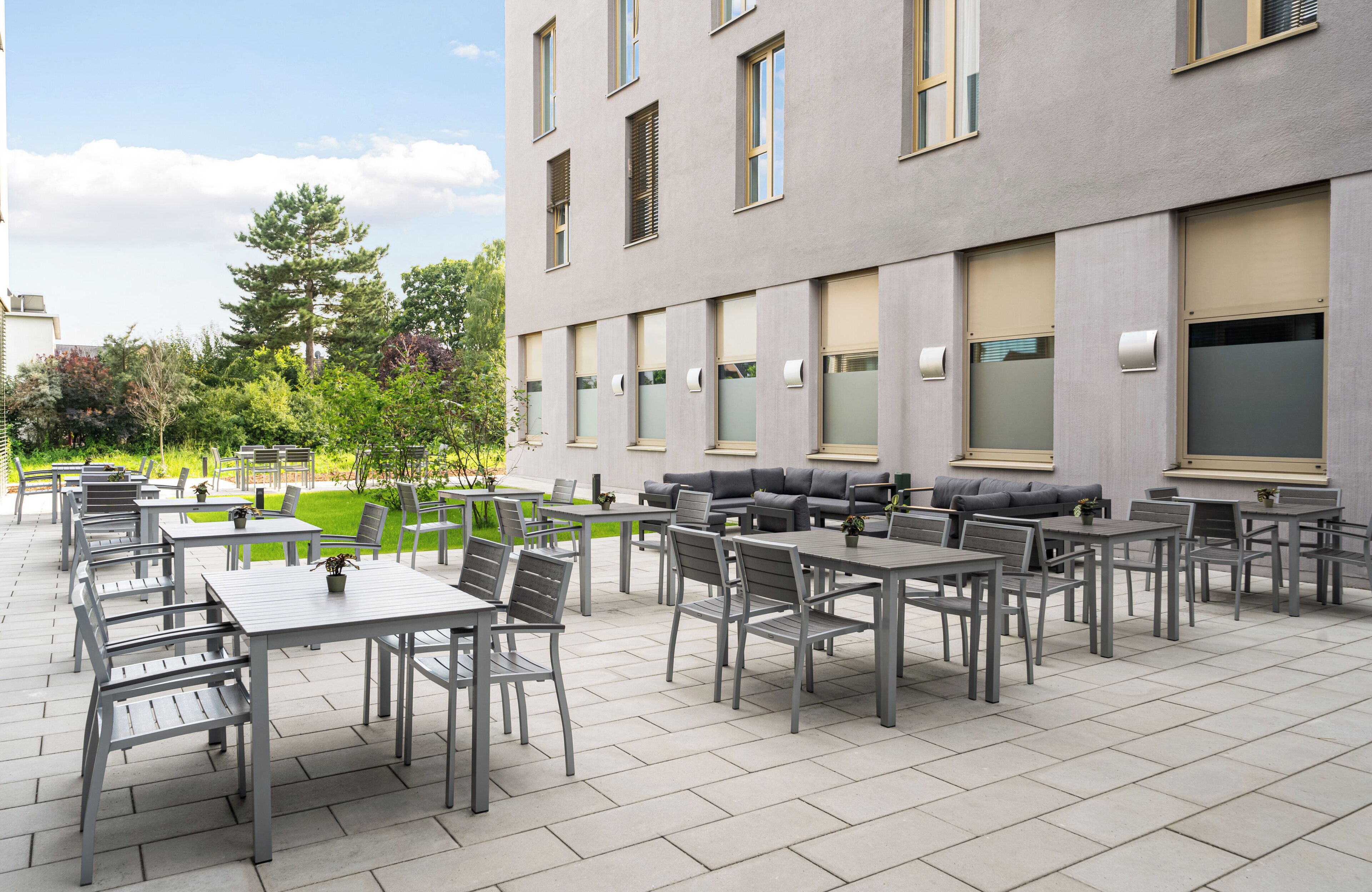 Ein Außenbereich mit einer Terrasse, mehreren Tischen und Stühlen, einer Sitzecke mit Sofas und einem Gebäude mit großen Fenstern und einem flachen Dach.