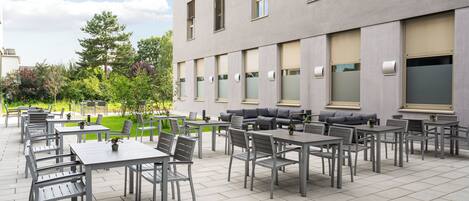 Ein Außenbereich mit einer Terrasse, mehreren Tischen und Stühlen, einer Sitzecke mit Sofas und einem Gebäude mit großen Fenstern und einem flachen Dach.