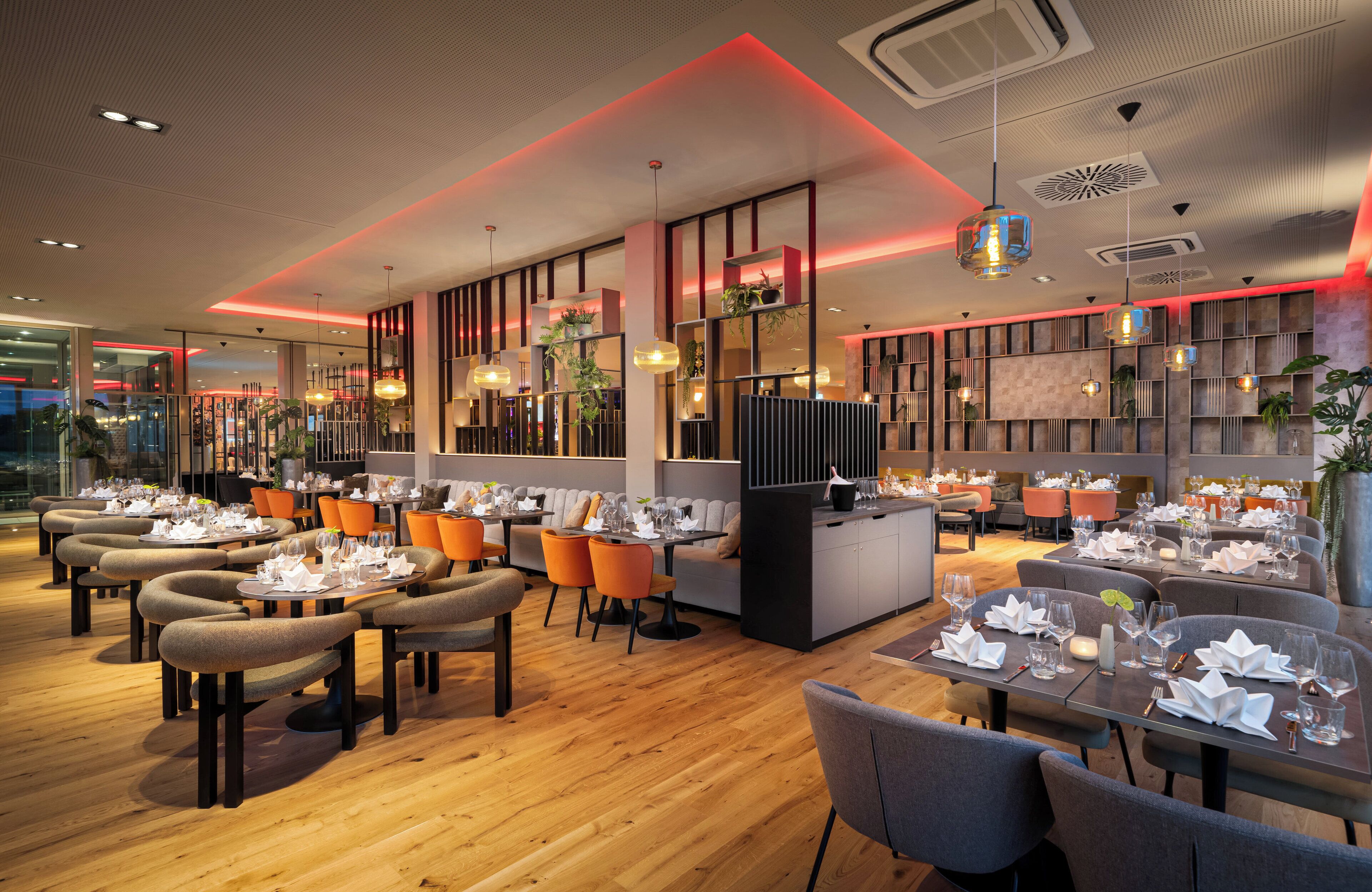 Un restaurante moderno con suelo de madera, sillas anaranjadas y grises, y mesas dispuestas con vajilla blanca.