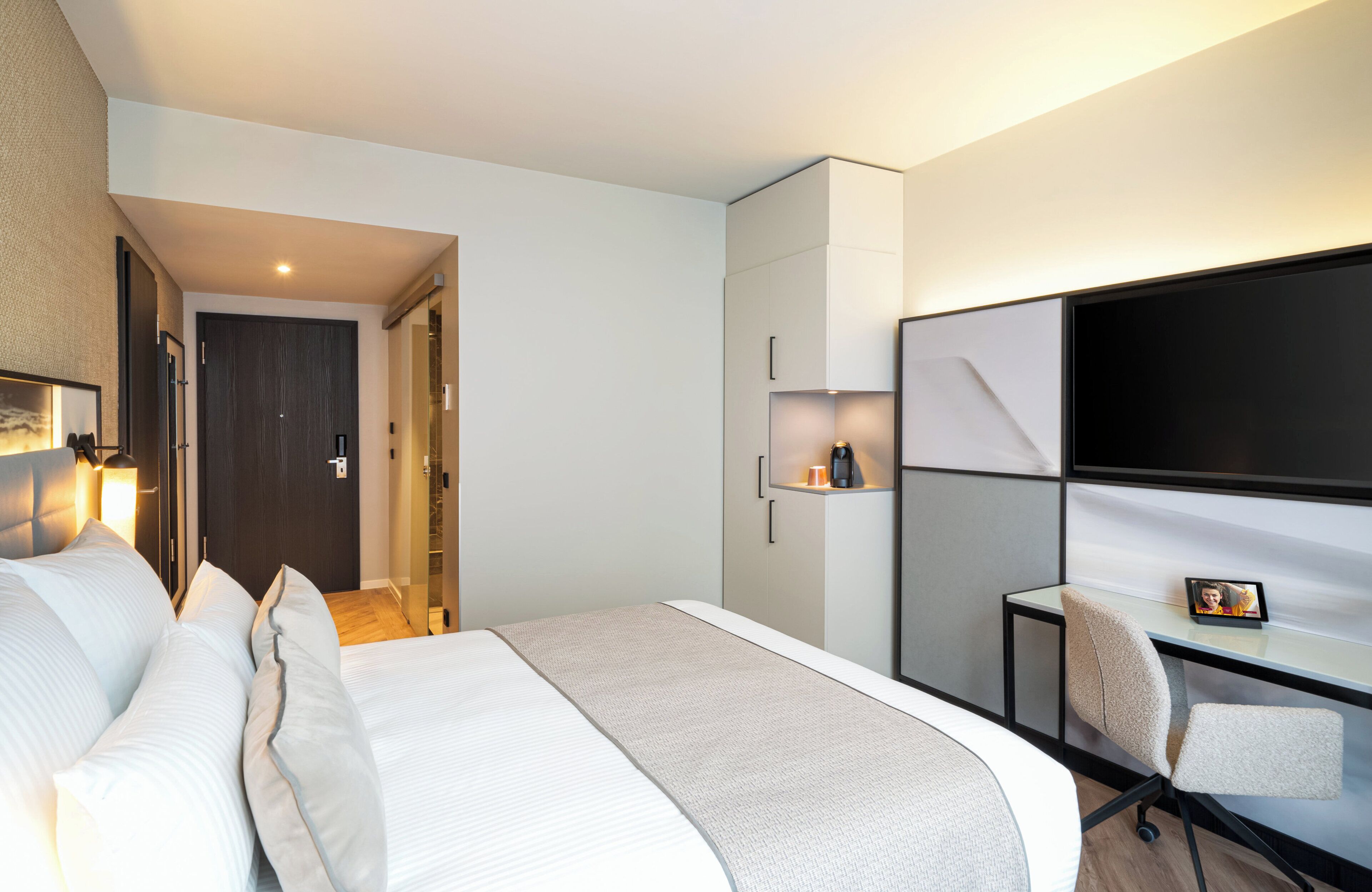 A modern hotel room with a large bed, a desk with a chair, a flat-screen TV, and a wardrobe.