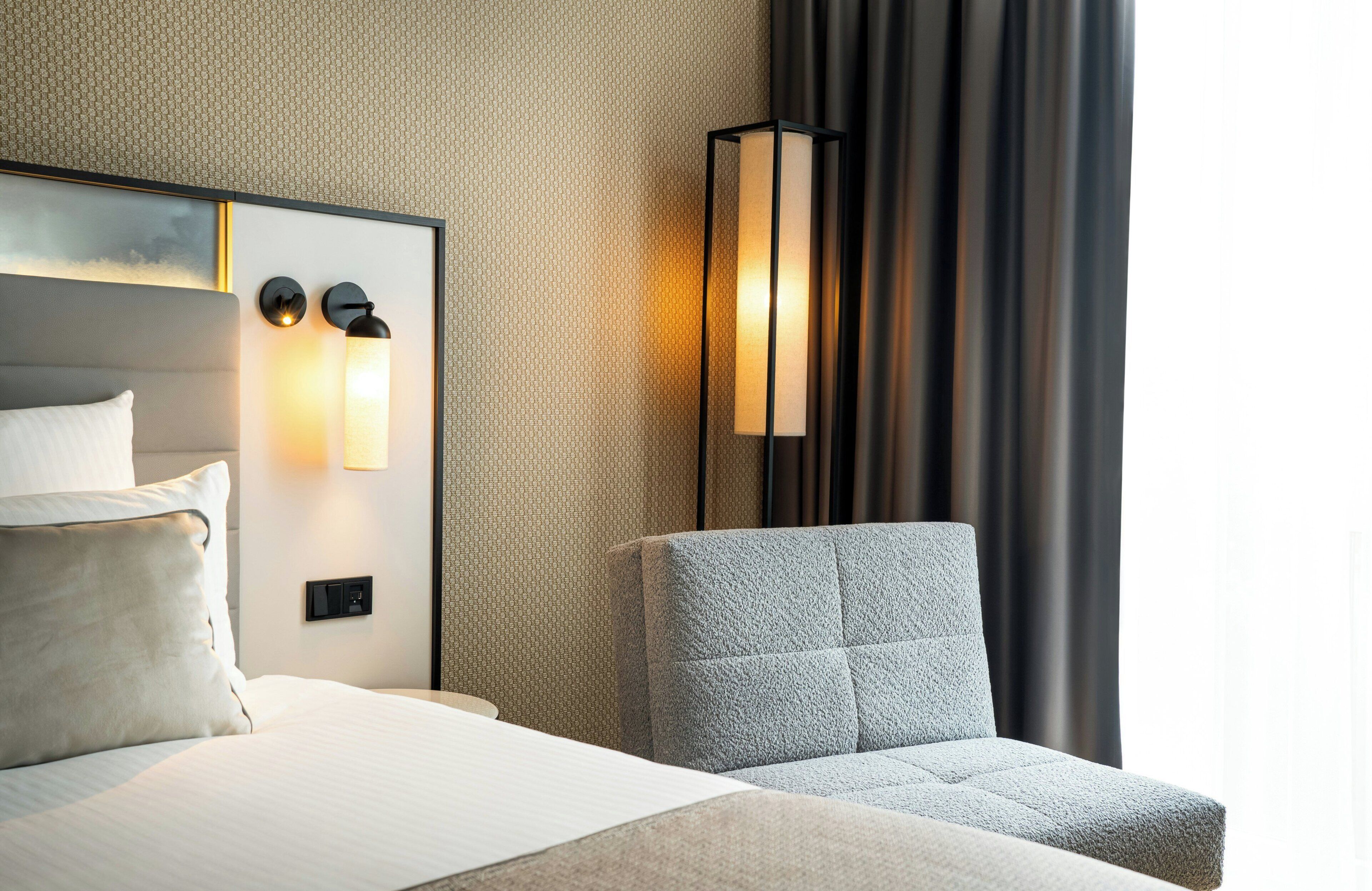 A hotel room with a bed, a nightstand, a wall-mounted lamp, and a chair.