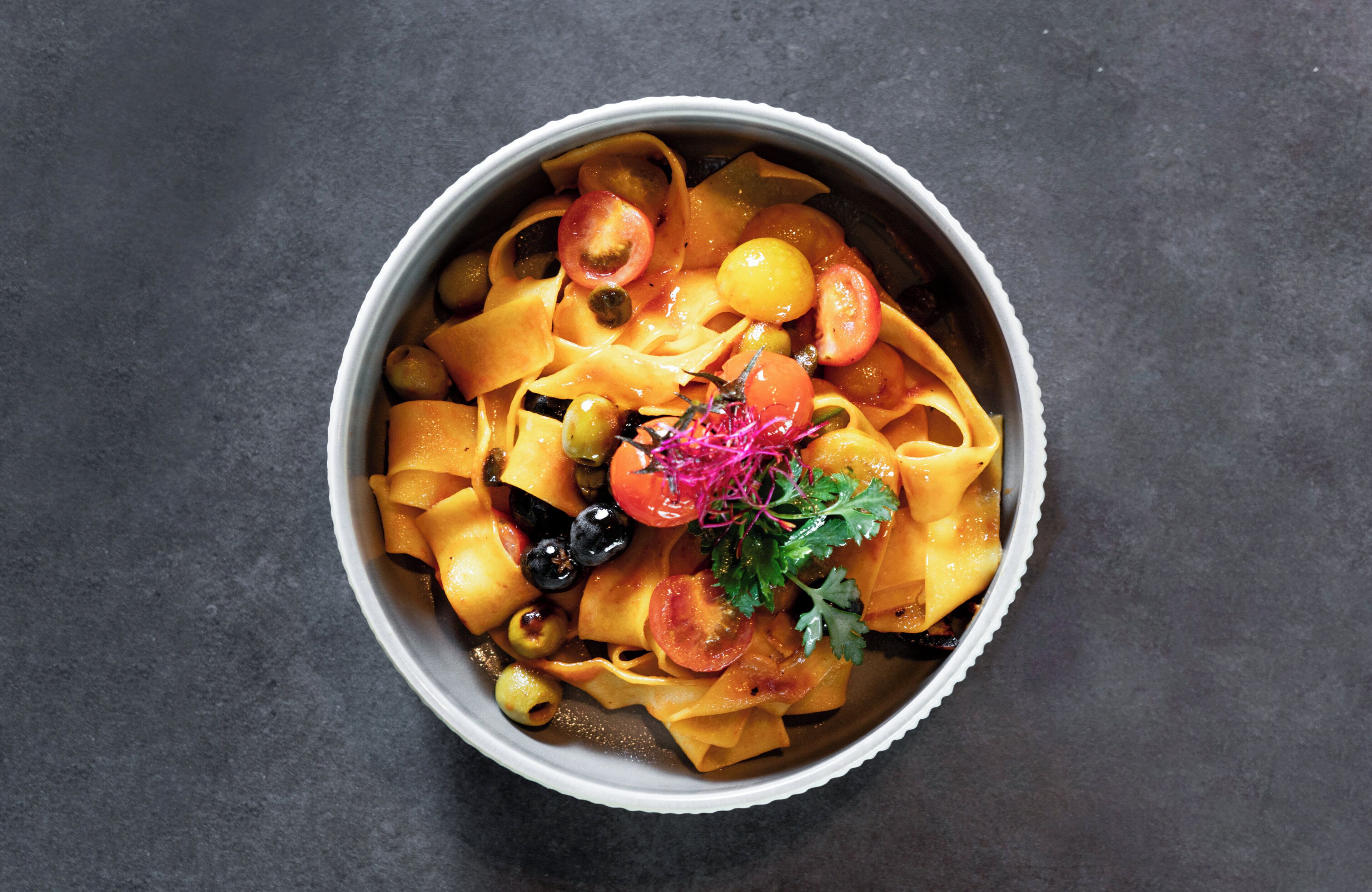 Pasta med tomater, oliven og pynt på en mørk overflade.