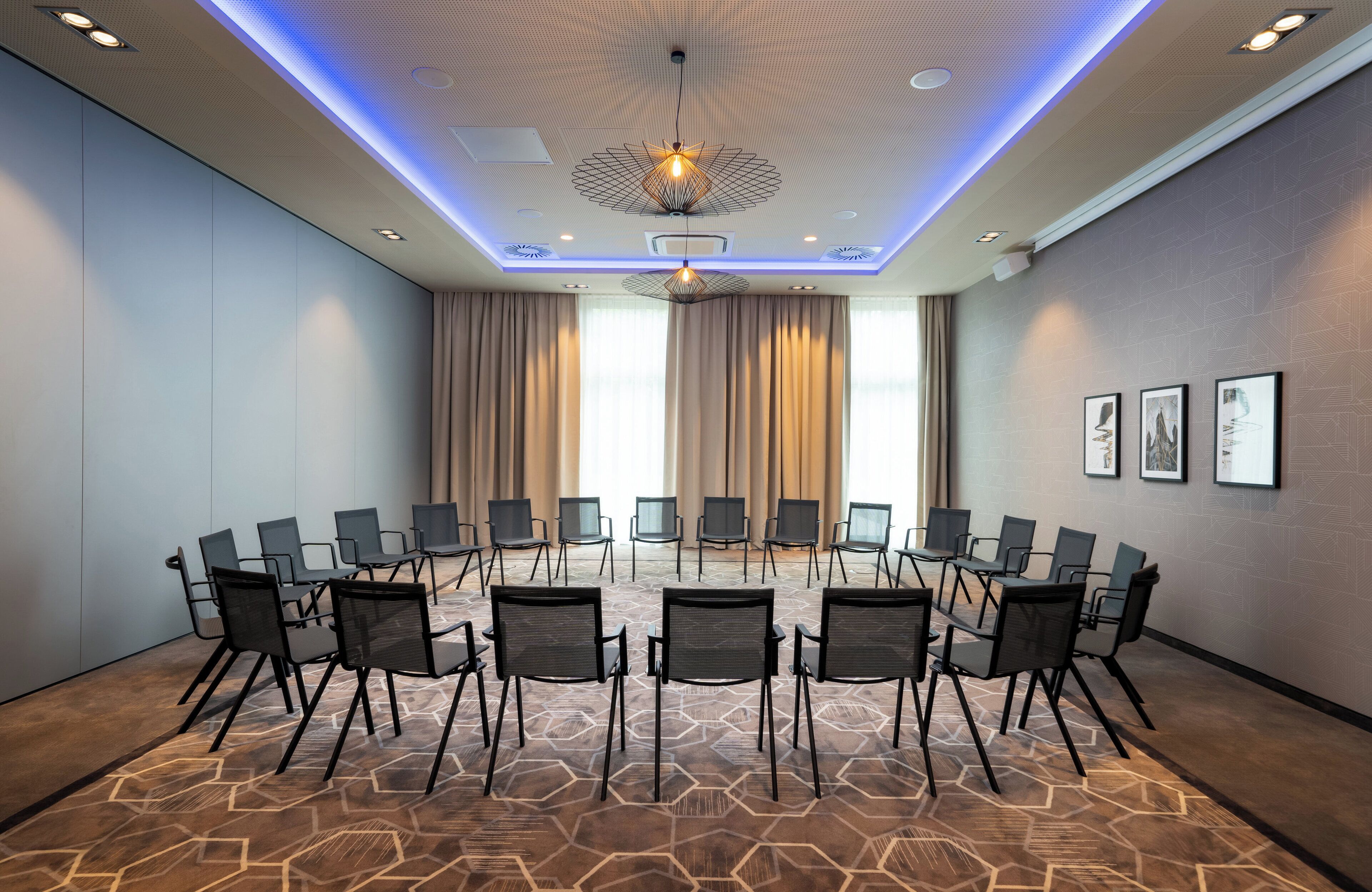 Una sala de conferencias con alfombra de estampado hexagonal, sillas negras dispuestas en círculo, y un techo moderno con iluminación empotrada y una lámpara central.