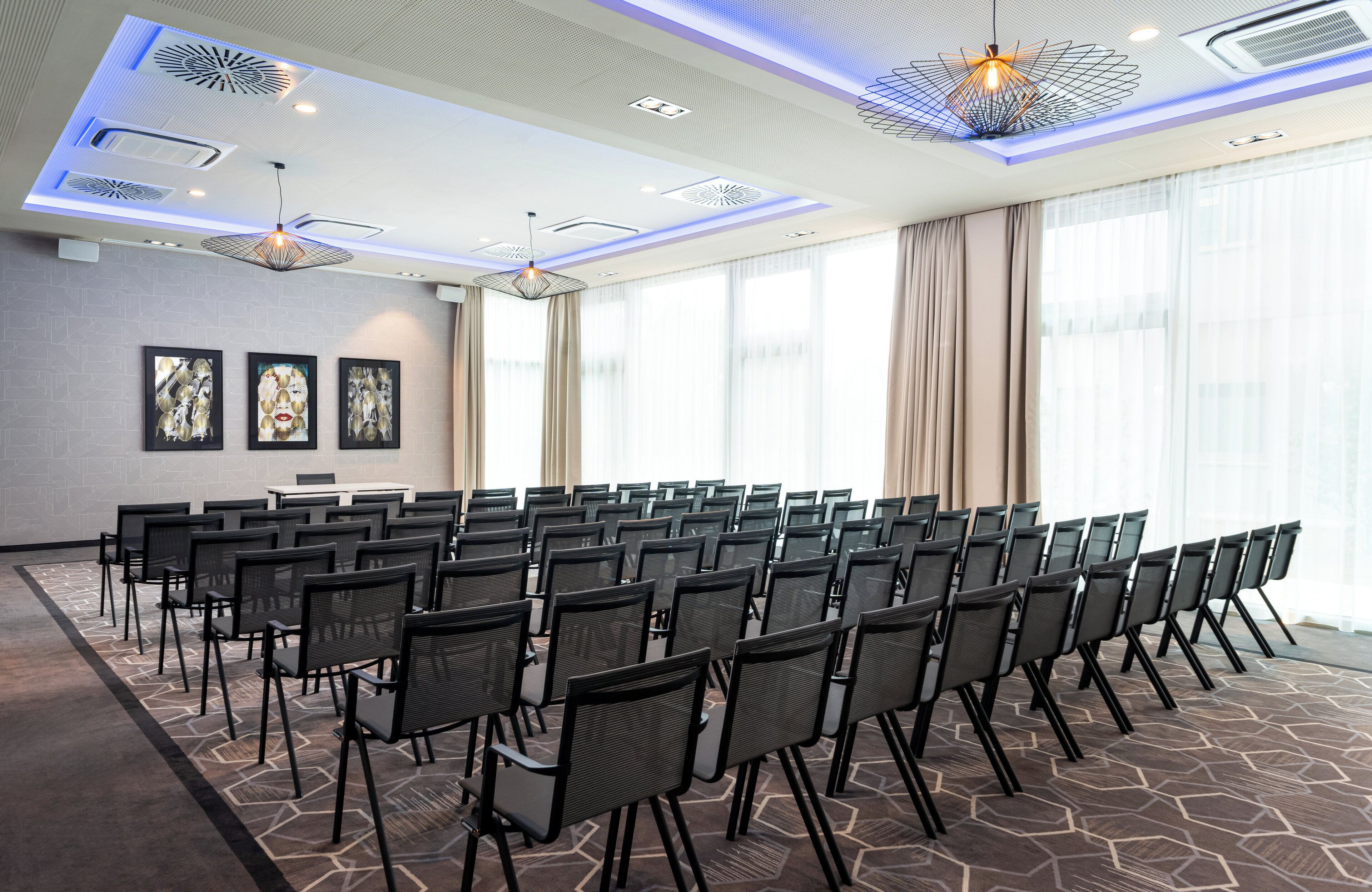Un salón de conferencias con filas de sillas, un ventanal con cortinas, e iluminación empotrada y detalles decorativos en el techo.