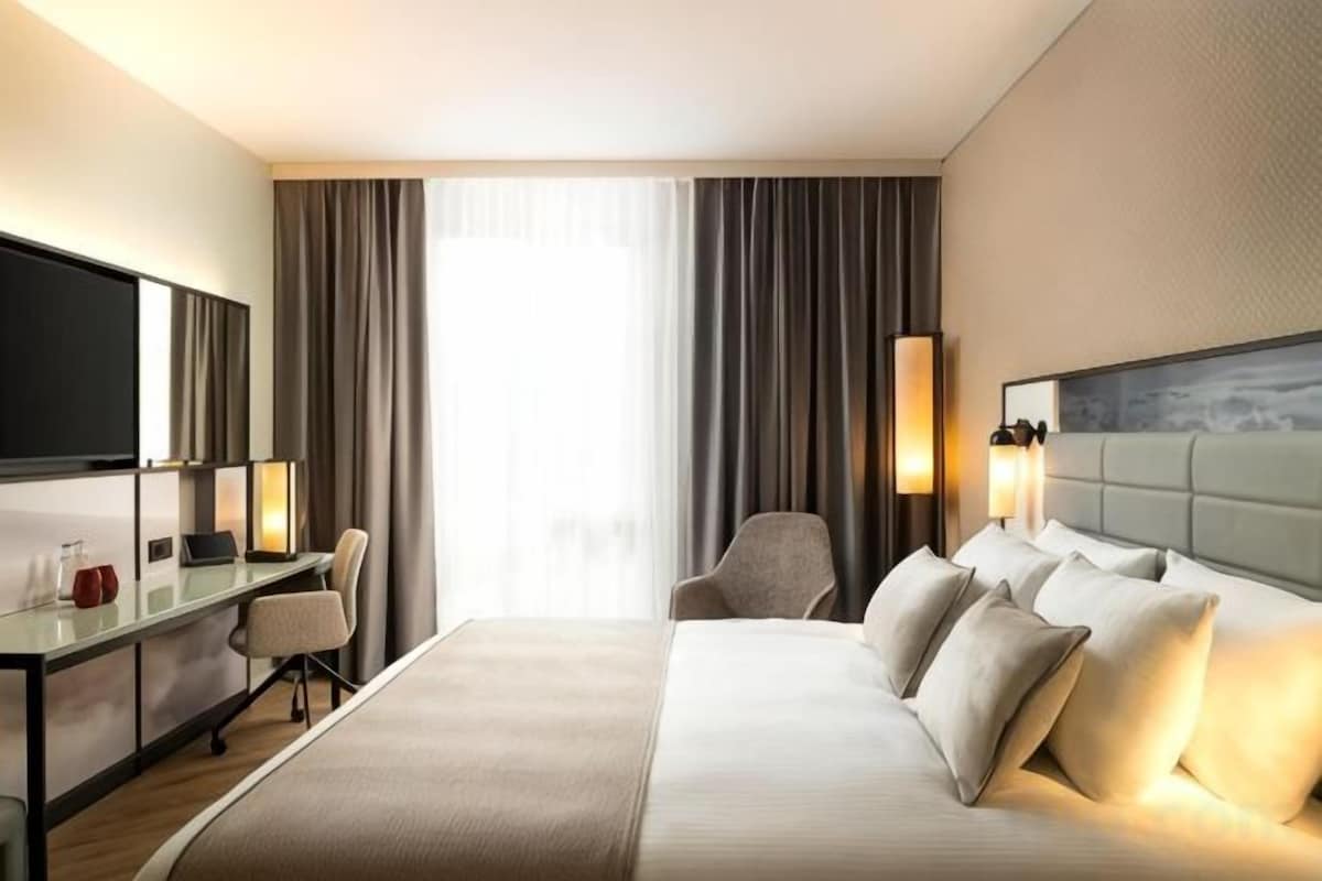 Habitación de hotel con cama, escritorio, silla, televisión y ventana con cortinas.
