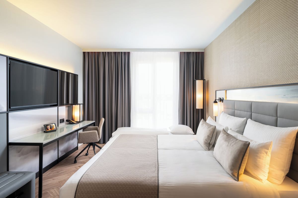 A modern hotel room with a large bed, a desk with a flat-screen TV, a chair, and a large window with curtains.