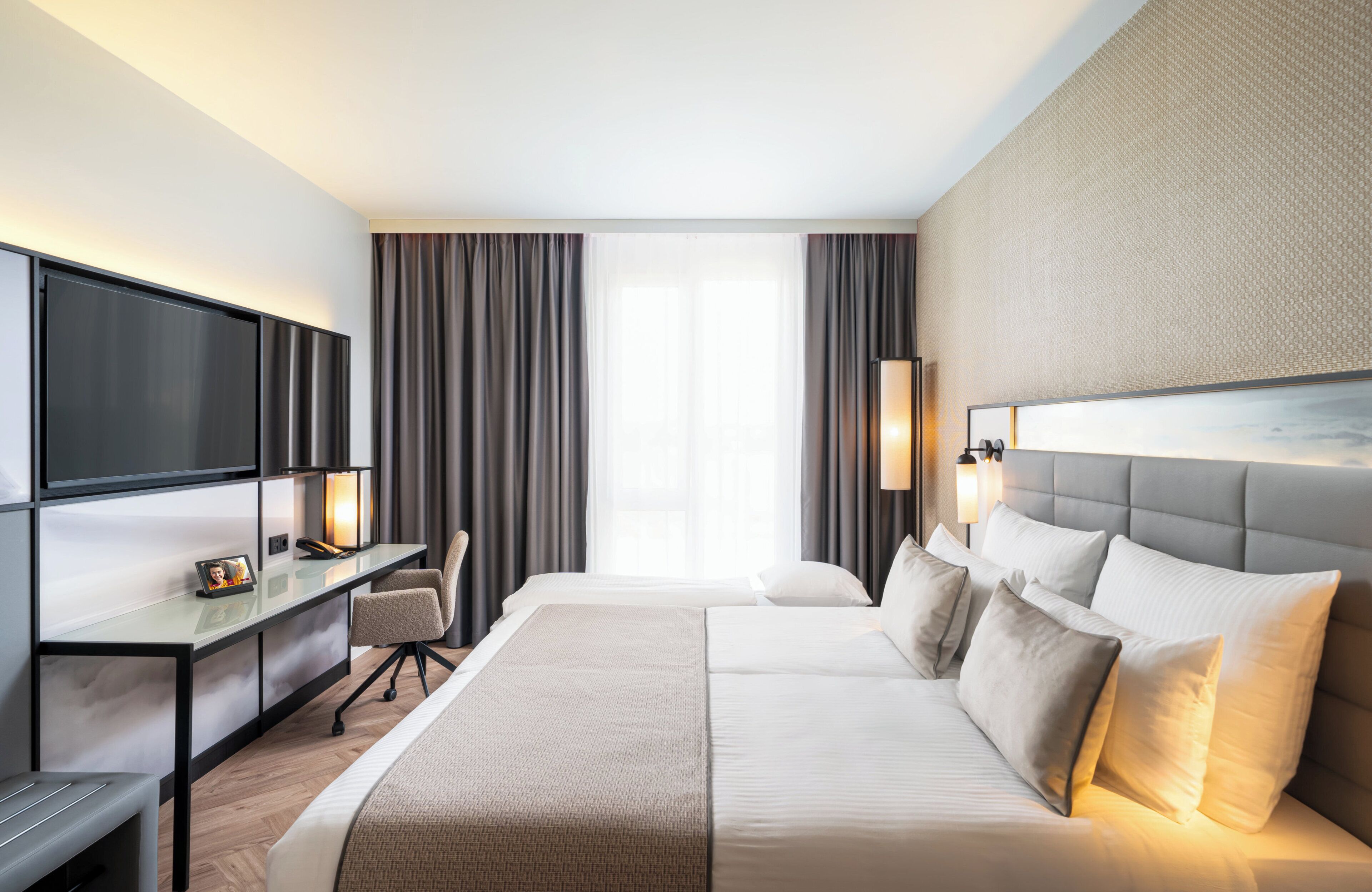 A modern hotel room with a large bed, a desk with a flat-screen TV, a chair, and a large window with curtains.