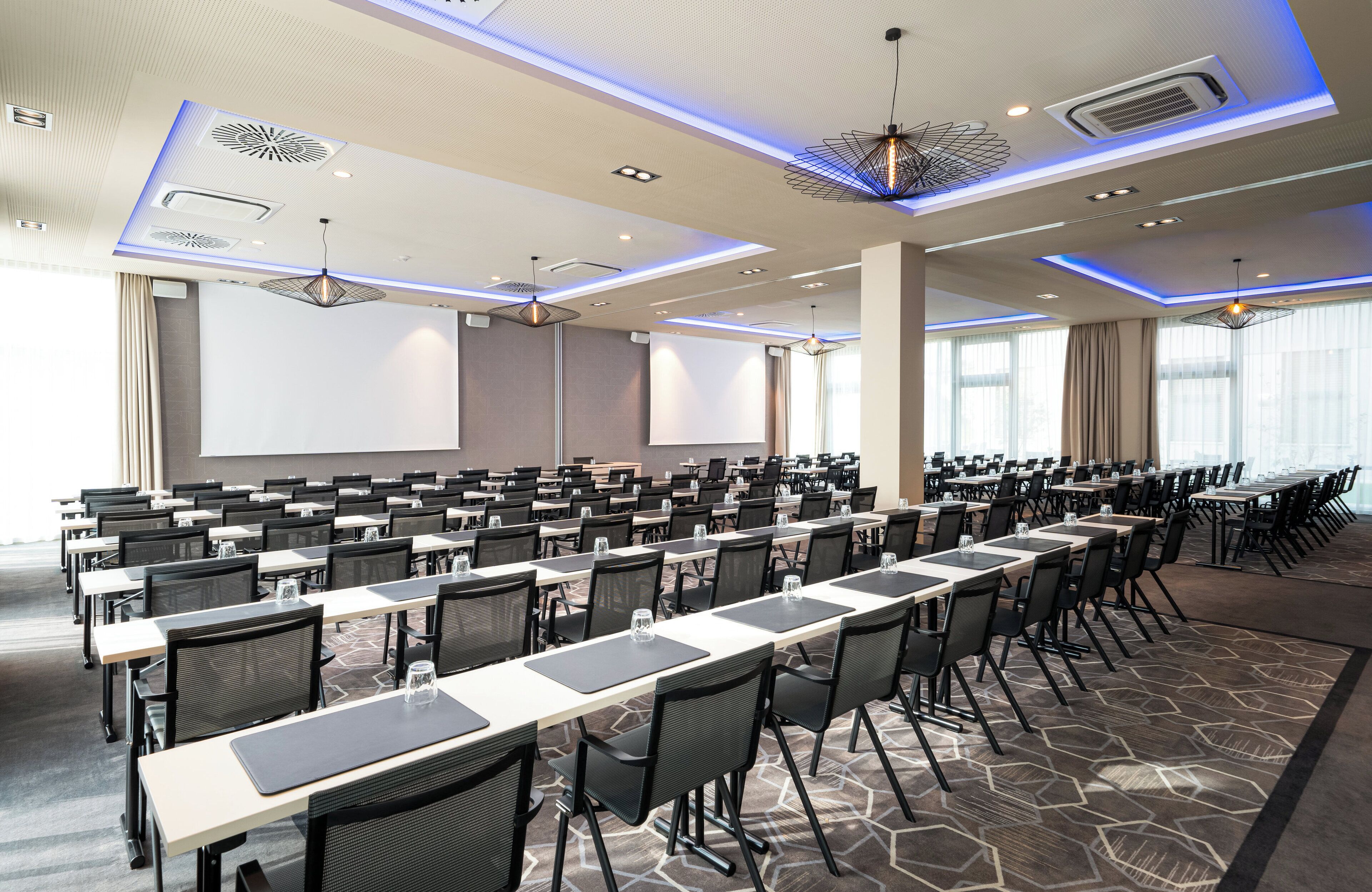 A conference room with rows of tables and chairs, large windows, and a projector screen.