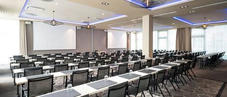 A conference room with rows of tables and chairs, large windows, and a projector screen.