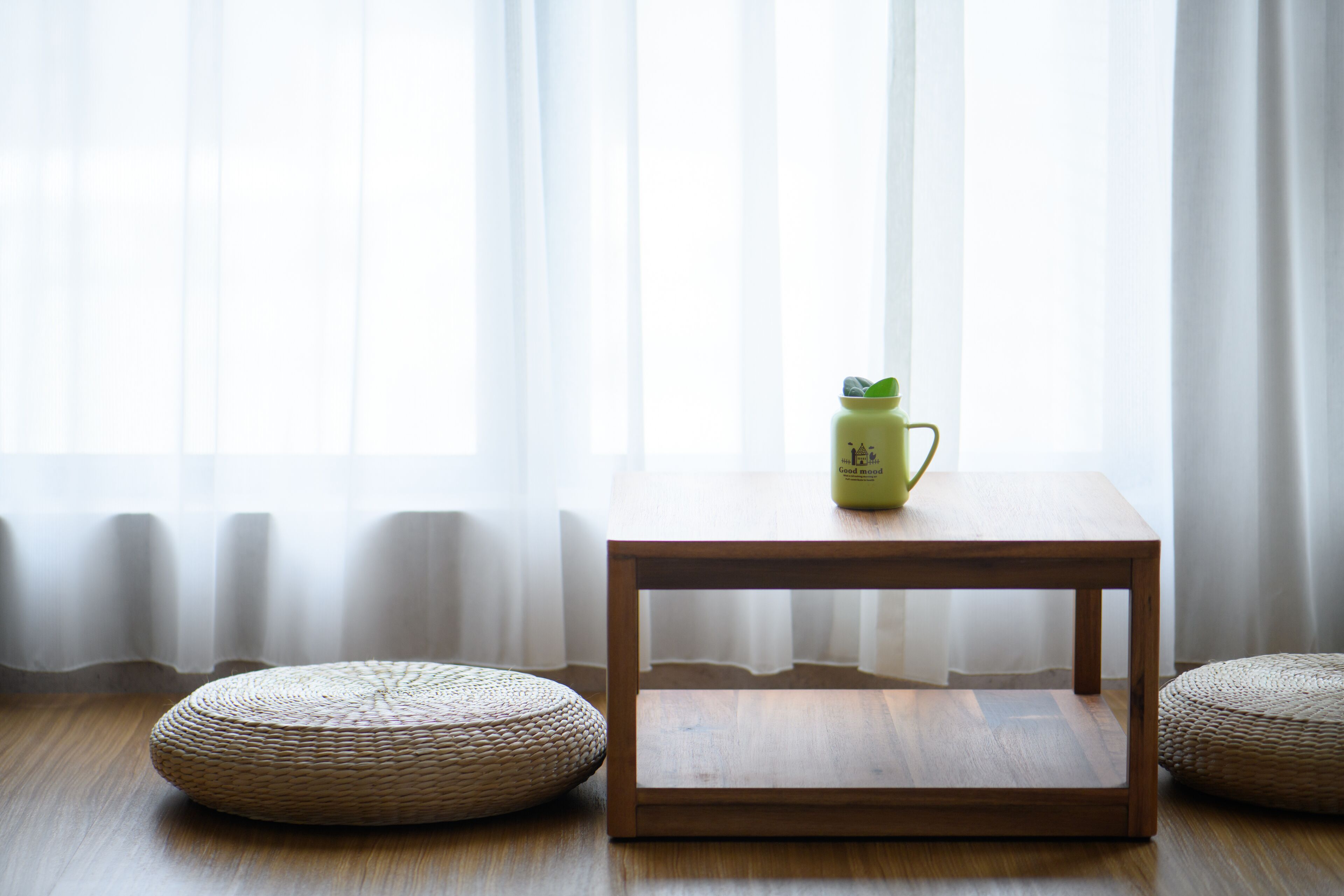 Una habitación con una mesa de madera, dos pufs tejidos y una botella verde sobre la mesa.