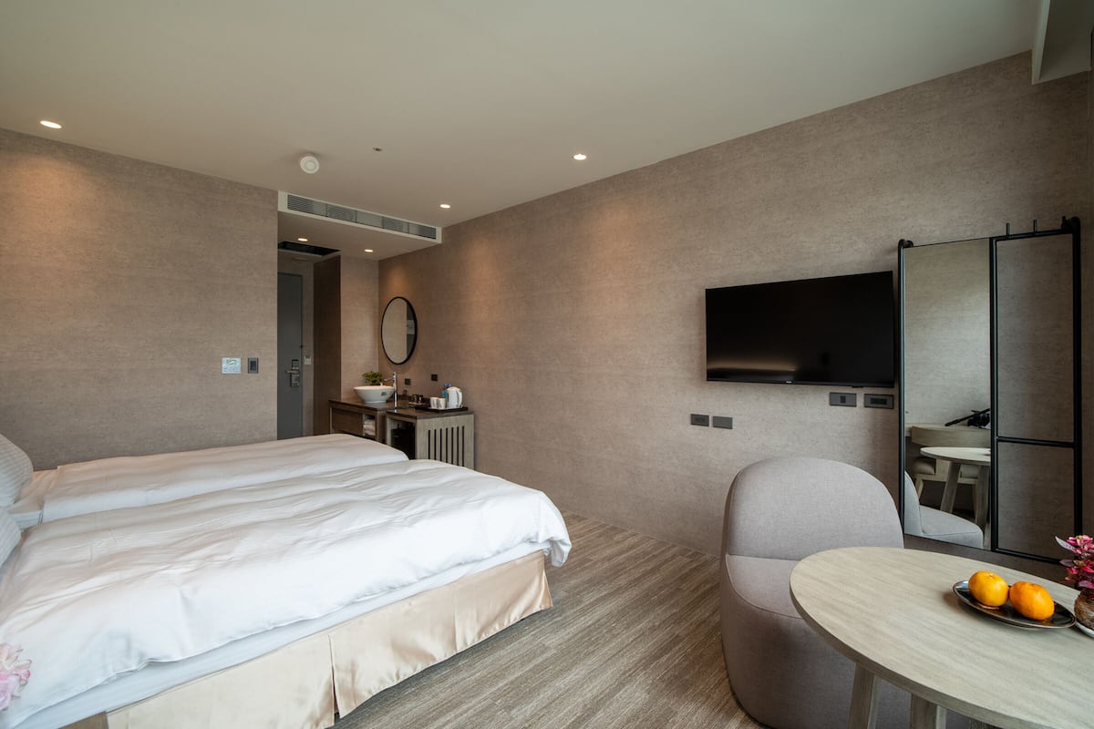 A modern hotel room with a large bed, a desk with a mirror, a flat-screen TV, and a small table with oranges and flowers.