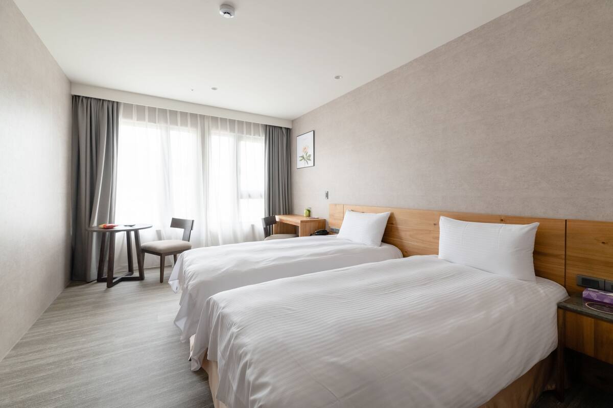 A hotel room with two beds, a desk, a chair, and a window with curtains.