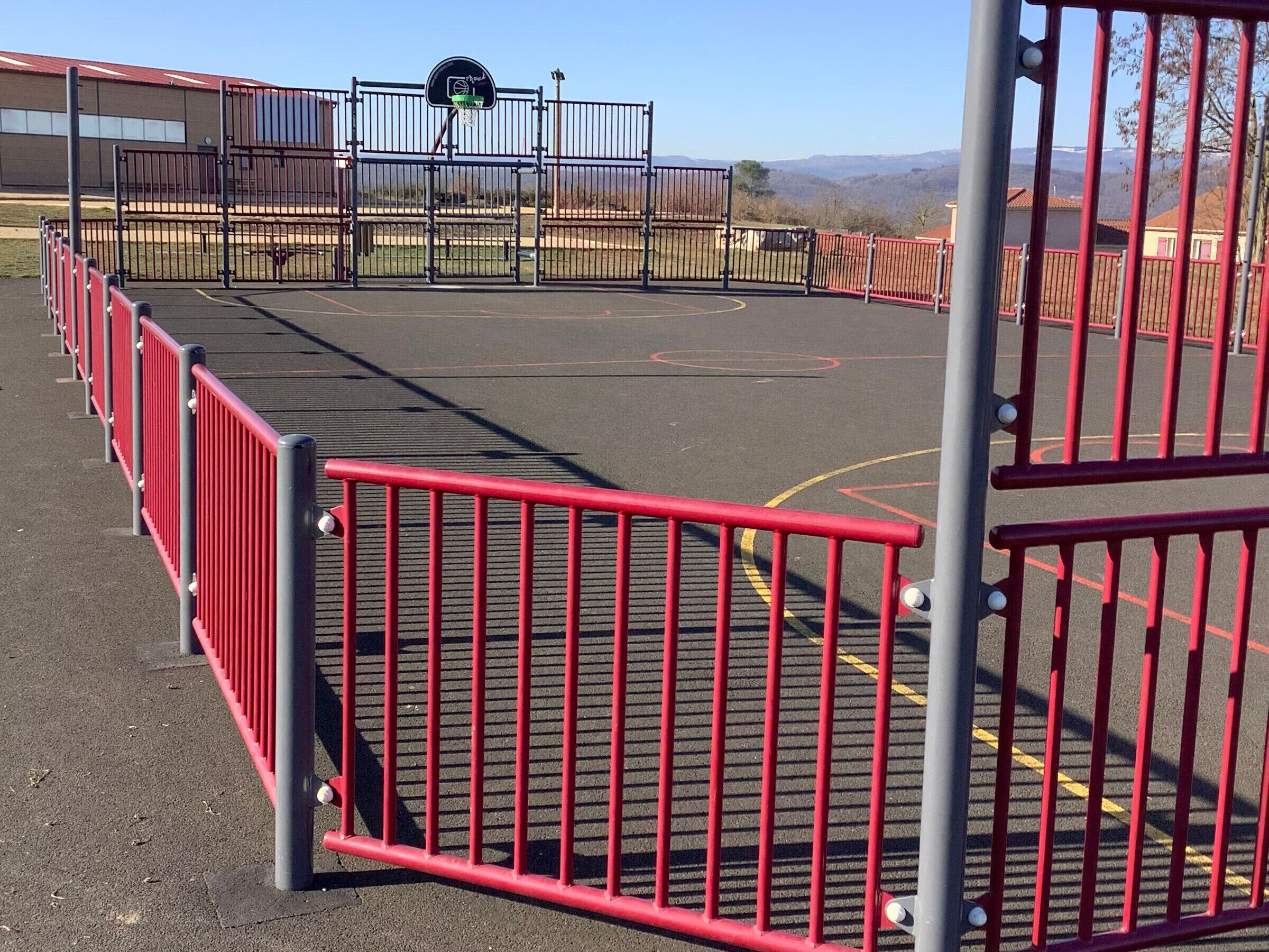 Un terrain de basket-ball avec des barrières rouges et noires, un panier de basket et un bâtiment en arrière-plan.
