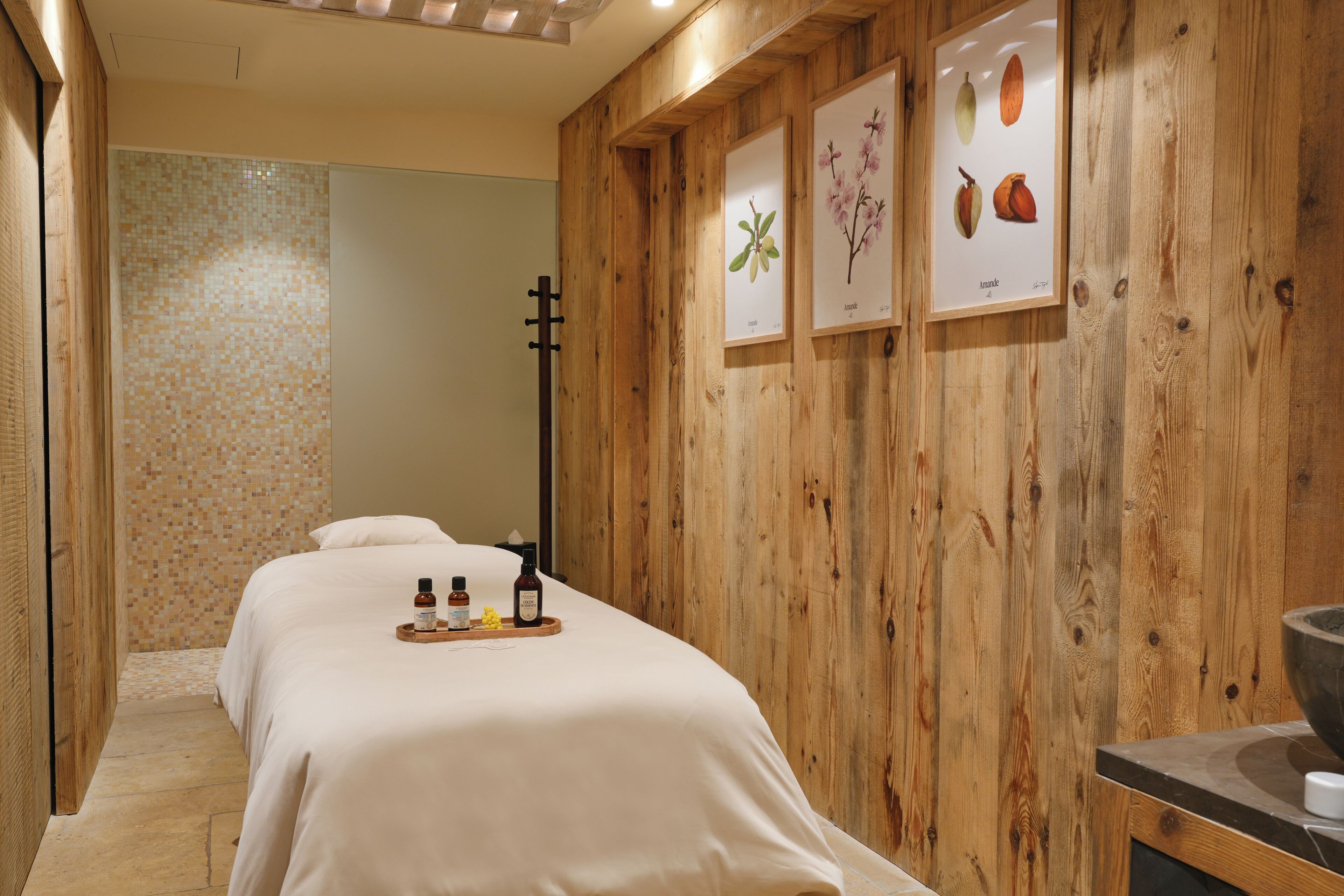 A spa room with wooden walls, two beds, a tray with bottles, and framed botanical prints.