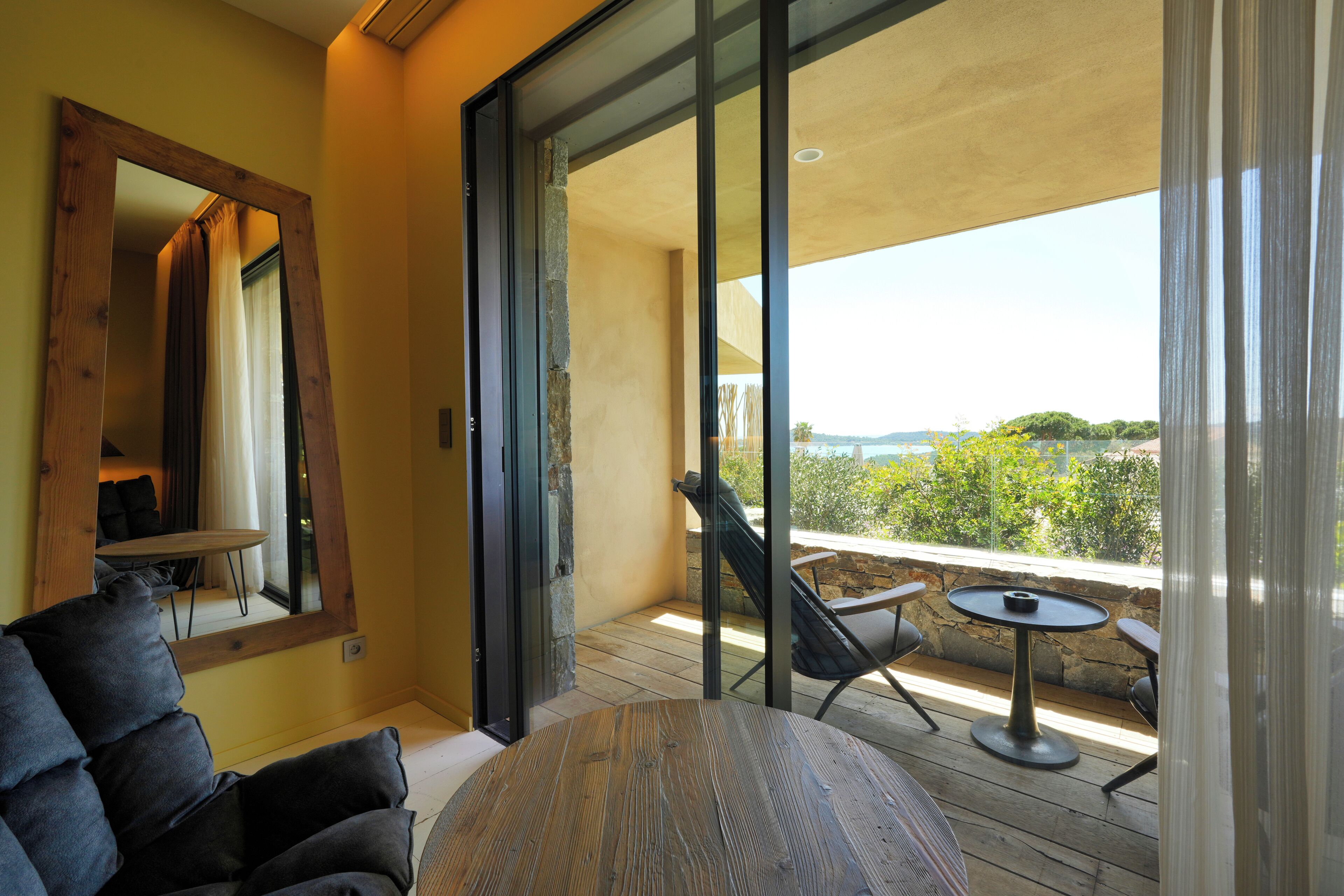 A modern living room with a large mirror, a round wooden table, and a balcony with a view of the countryside.