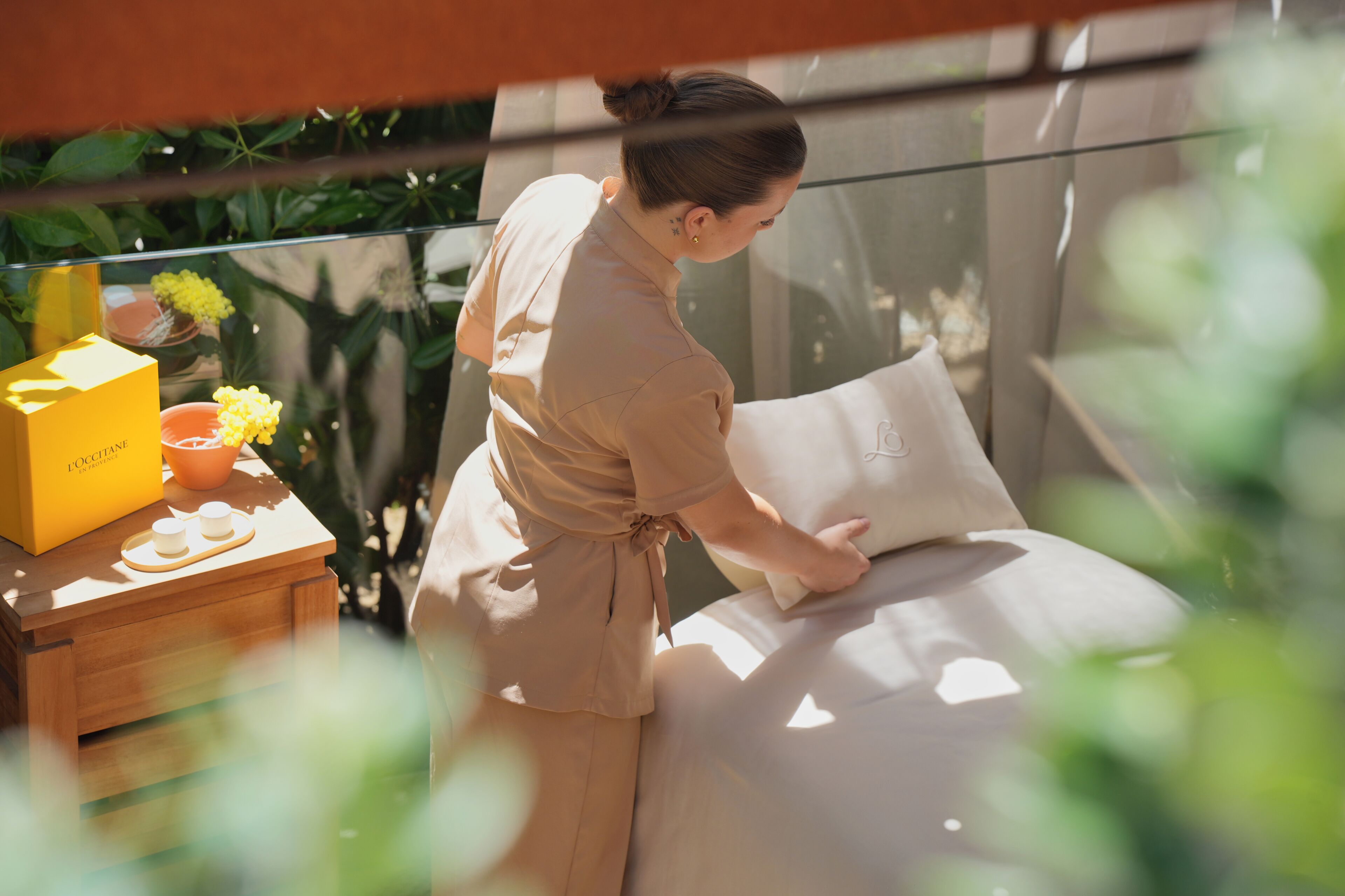 A person adjusting a bed in a room with a glass partition, a wooden nightstand, and a yellow box.
