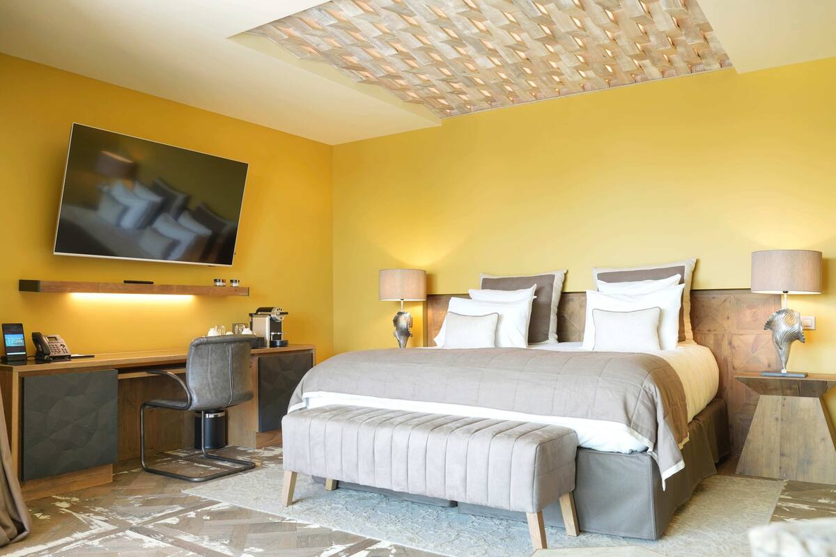 A hotel room with a bed, bedside lamps, a desk with a chair, a TV mounted on the wall, and a patterned rug.