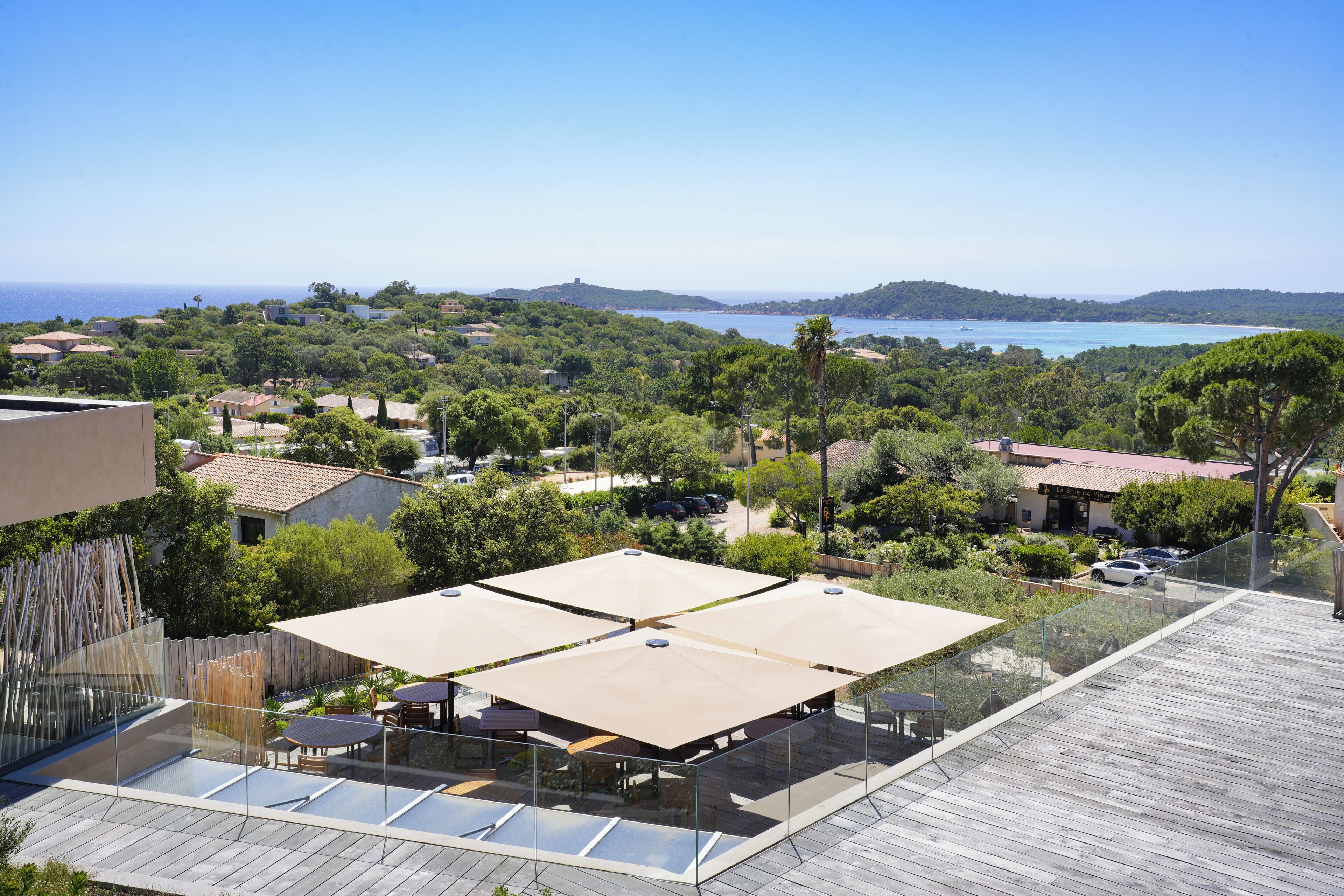 A rooftop terrace with glass railings, outdoor seating, and a view of the sea and hills.