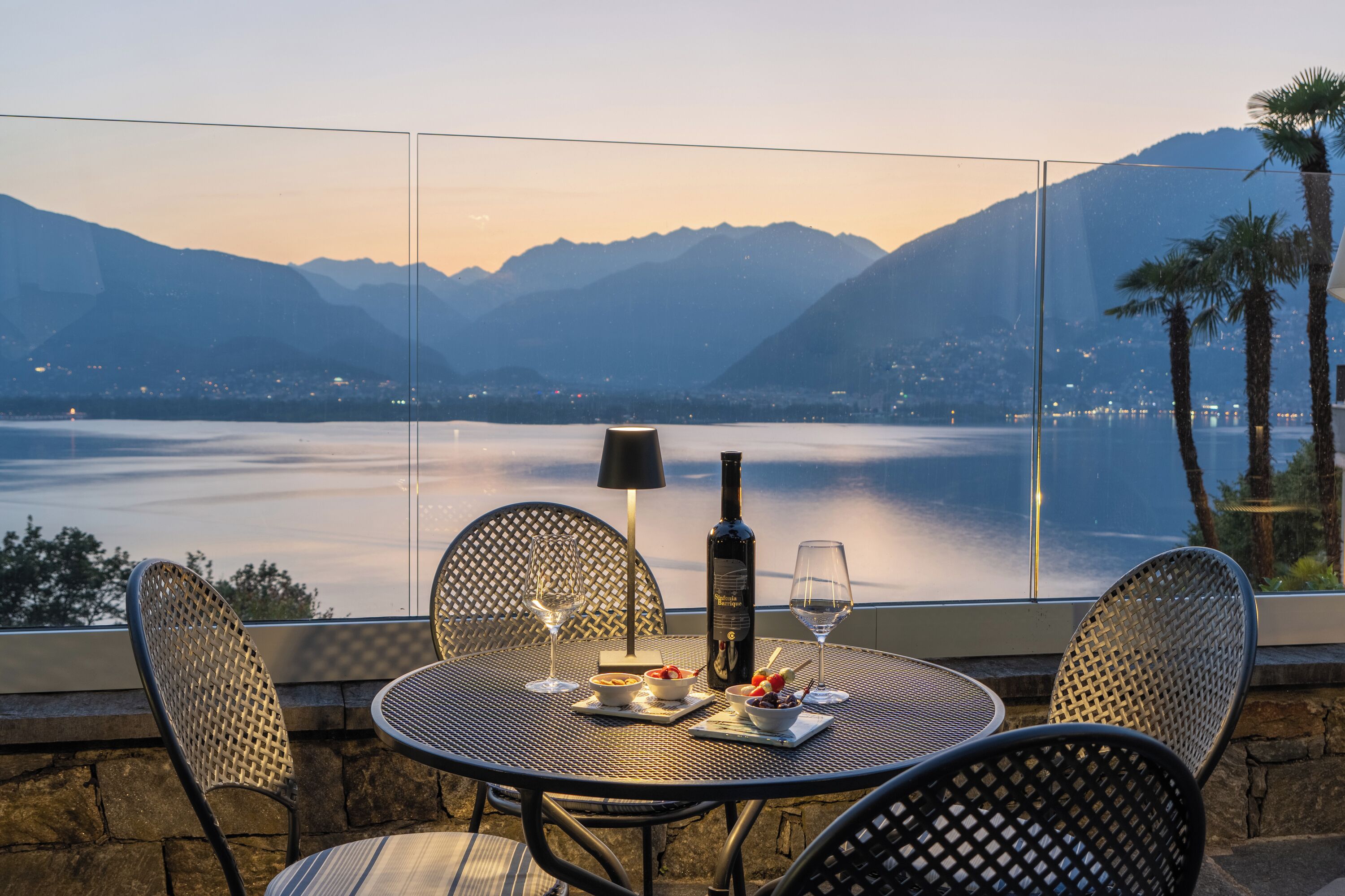 A table with chairs, a bottle, wine glasses, and a bowl of fruit, overlooking a lake with mountains in the background.