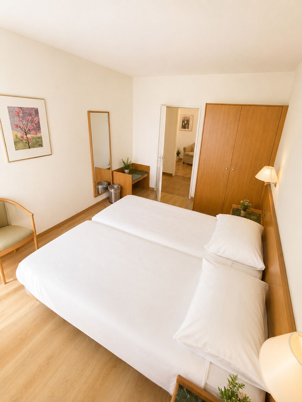 Habitación de hotel con una cama grande, un armario de madera, una mesita de noche con lámpara, una silla y un cuadro enmarcado en la pared.