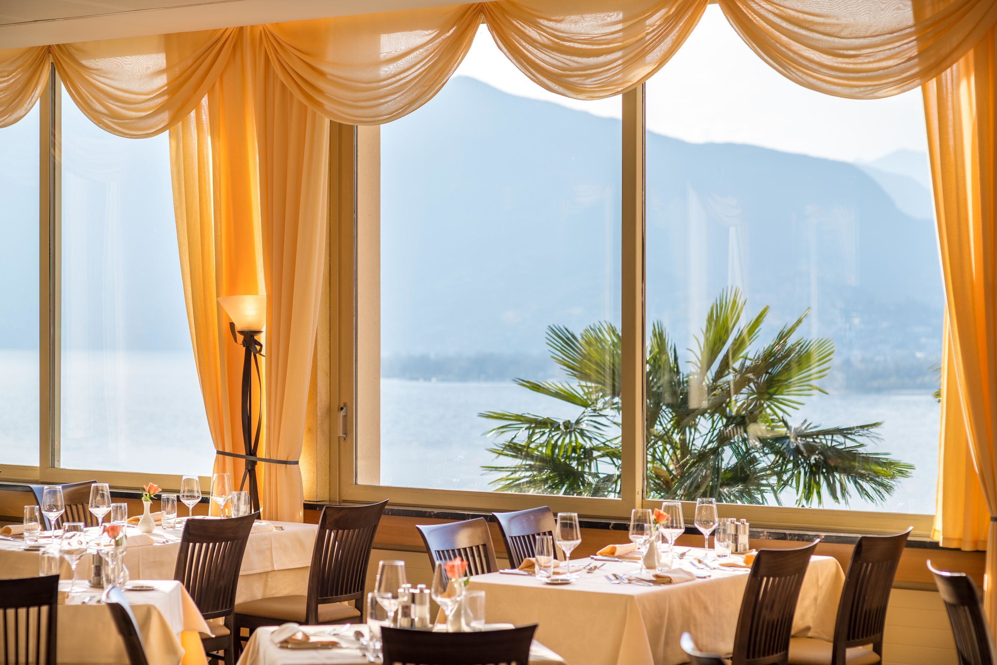 Un comedor con mesas dispuestas para una comida, ventanas amplias con cortinas y vistas a un cuerpo de agua y montañas.