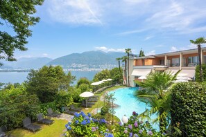 A pool area with lounge chairs and umbrellas, surrounded by lush greenery and a view of the sea and mountains. - Bellavista Swiss Quality Hotel    (Gambarogno)