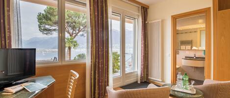 A hotel room with a TV, a desk, a chair, a sofa, and a view of the sea and mountains.