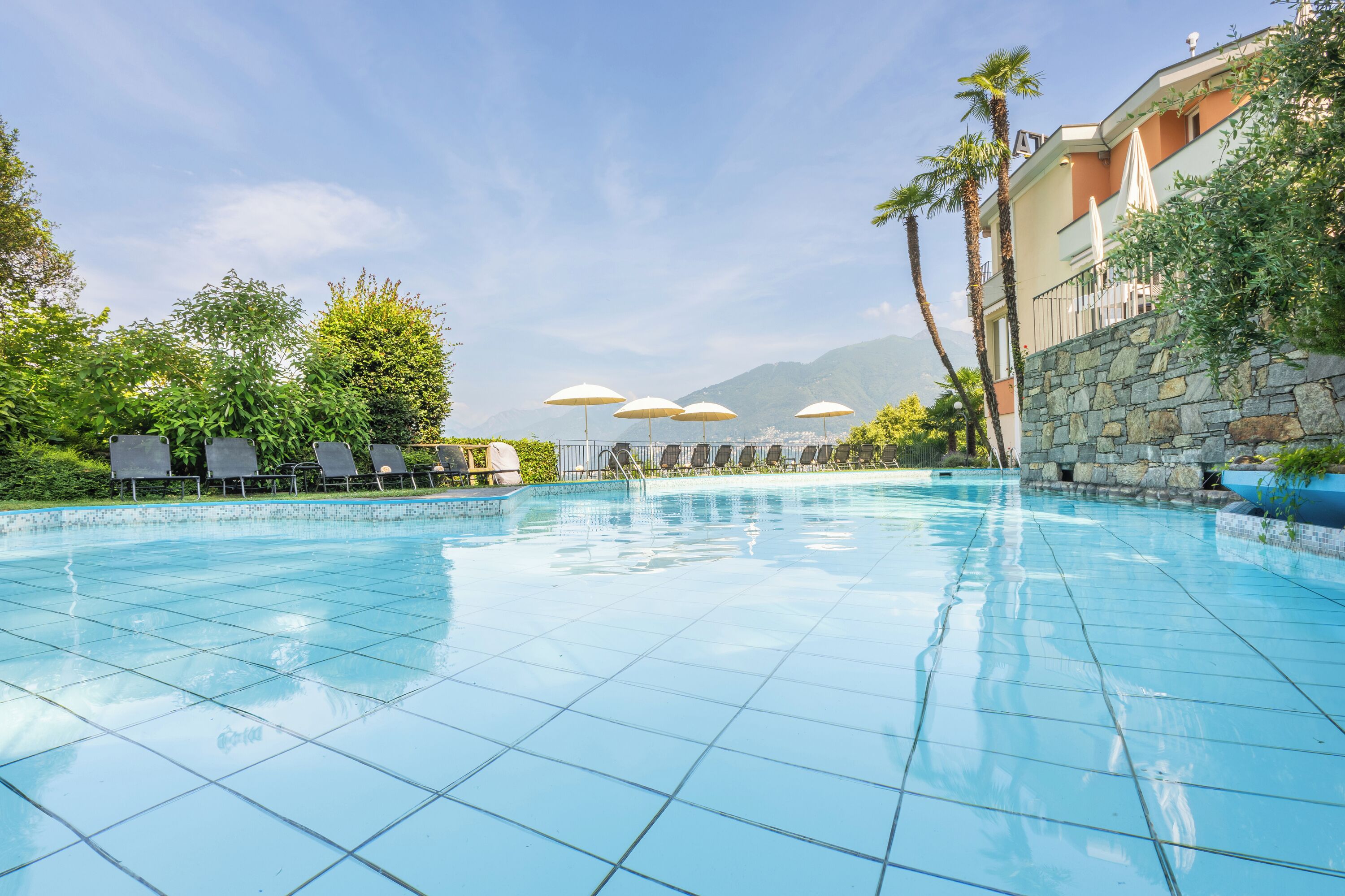 Ein klares, blaues Schwimmbecken mit Fliesenboden, umgeben von Liegestühlen und Sonnenschirmen, vor einer Kulisse aus Grünpflanzen und Bergen.