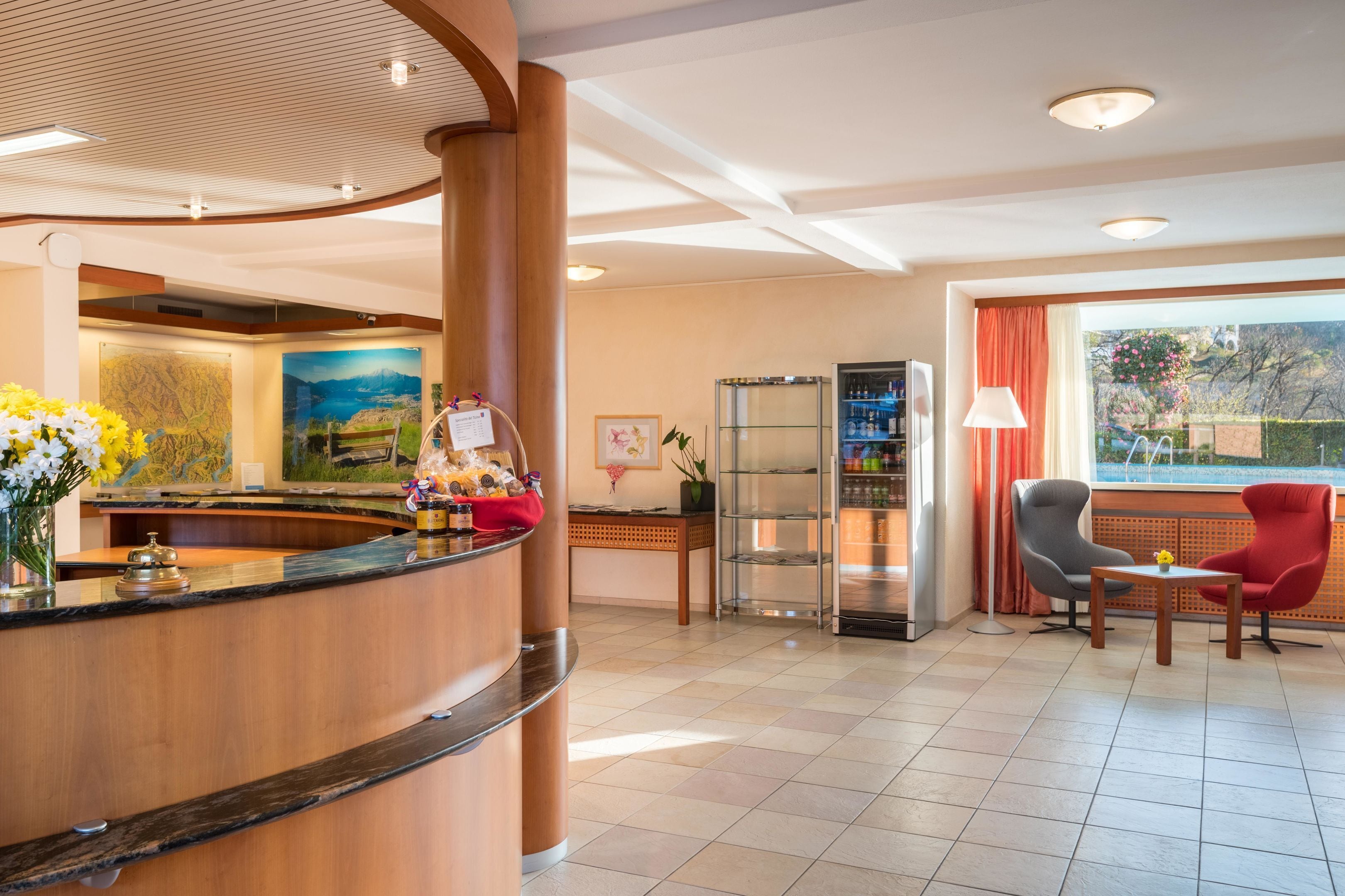 A hotel reception area with a wooden reception desk, a glass display case, a seating area with two chairs, and a large window offering a view of the outdoors.