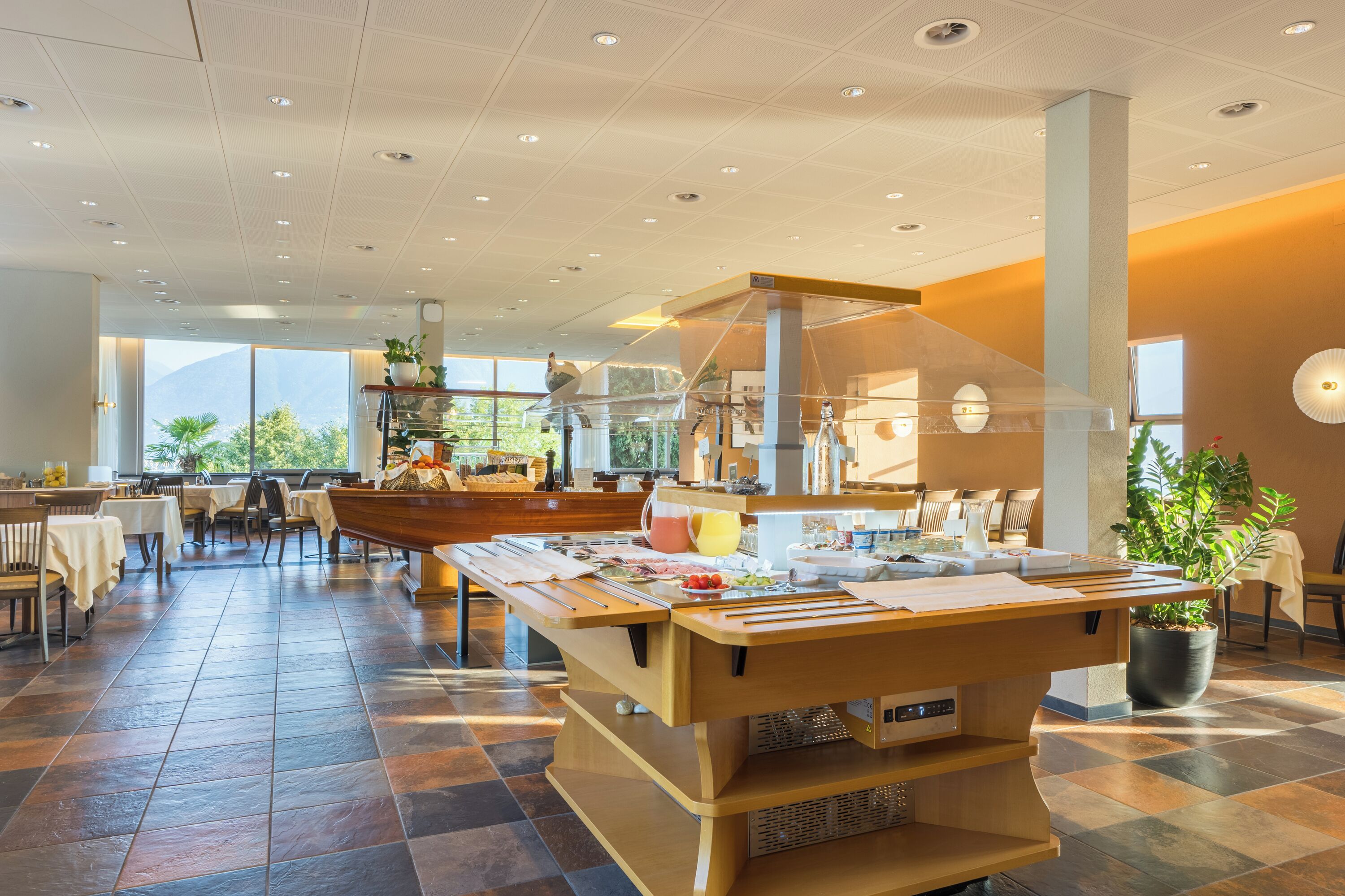 A buffet setup with a variety of food items, a seating area with tables and chairs, and a view of the outdoors through large windows.