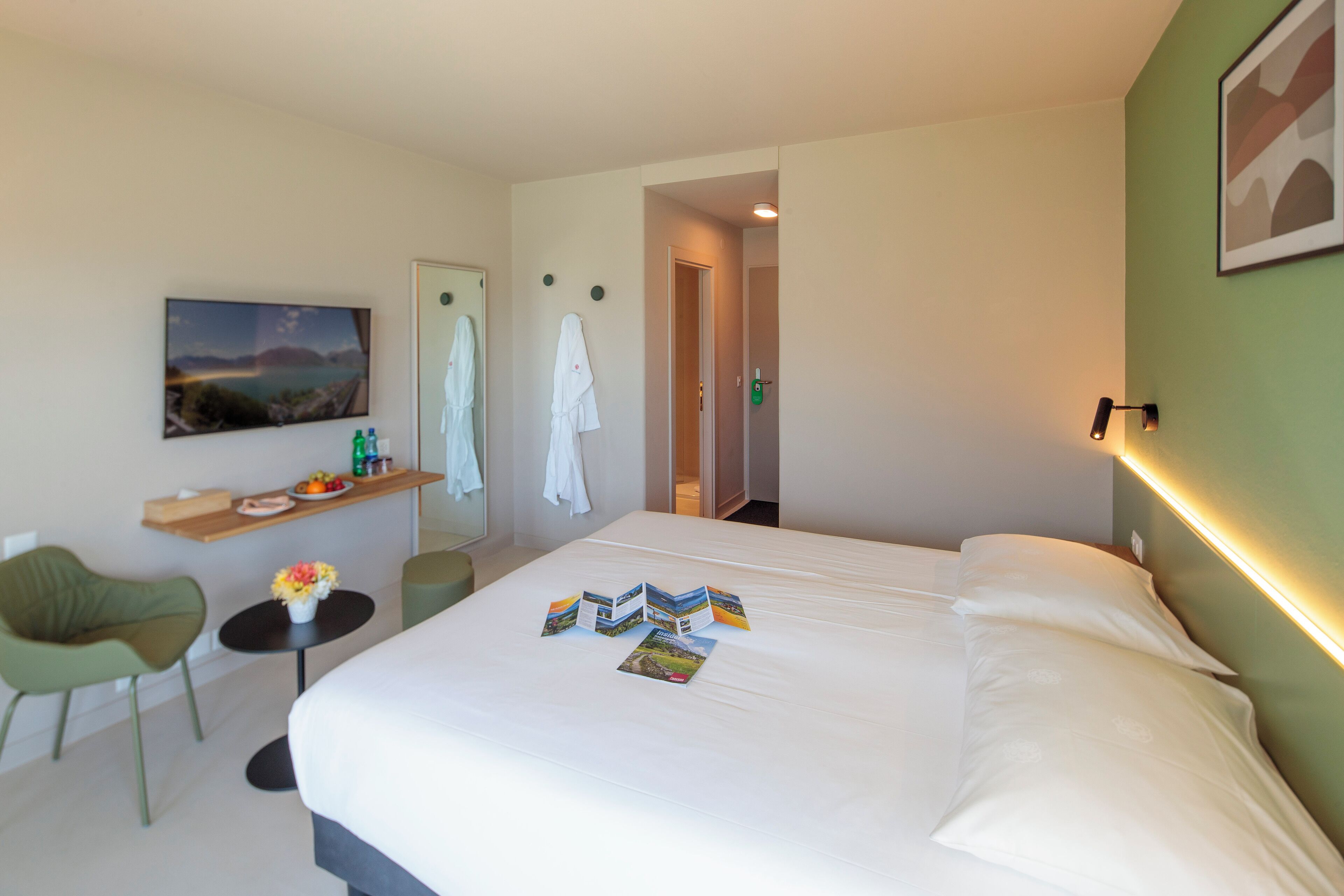 Habitación de hotel con cama, escritorio con silla, pequeña mesa con un florero y estante en la pared con artículos de aseo.