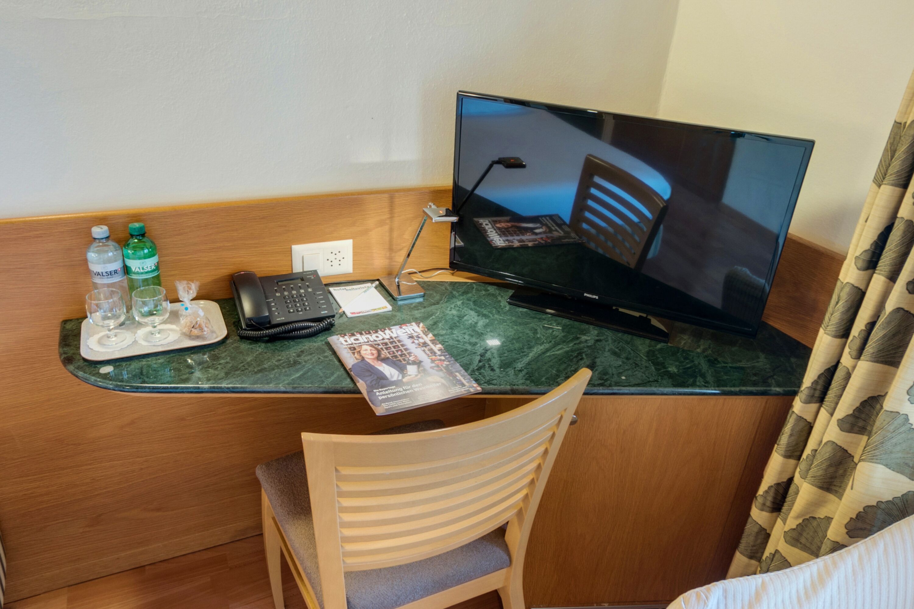 Ein Schreibtisch im Hotelzimmer mit einem Flachbildfernseher, einem Telefon, einer Zeitschrift und einem Tablett mit Gläsern und Wasserflaschen.