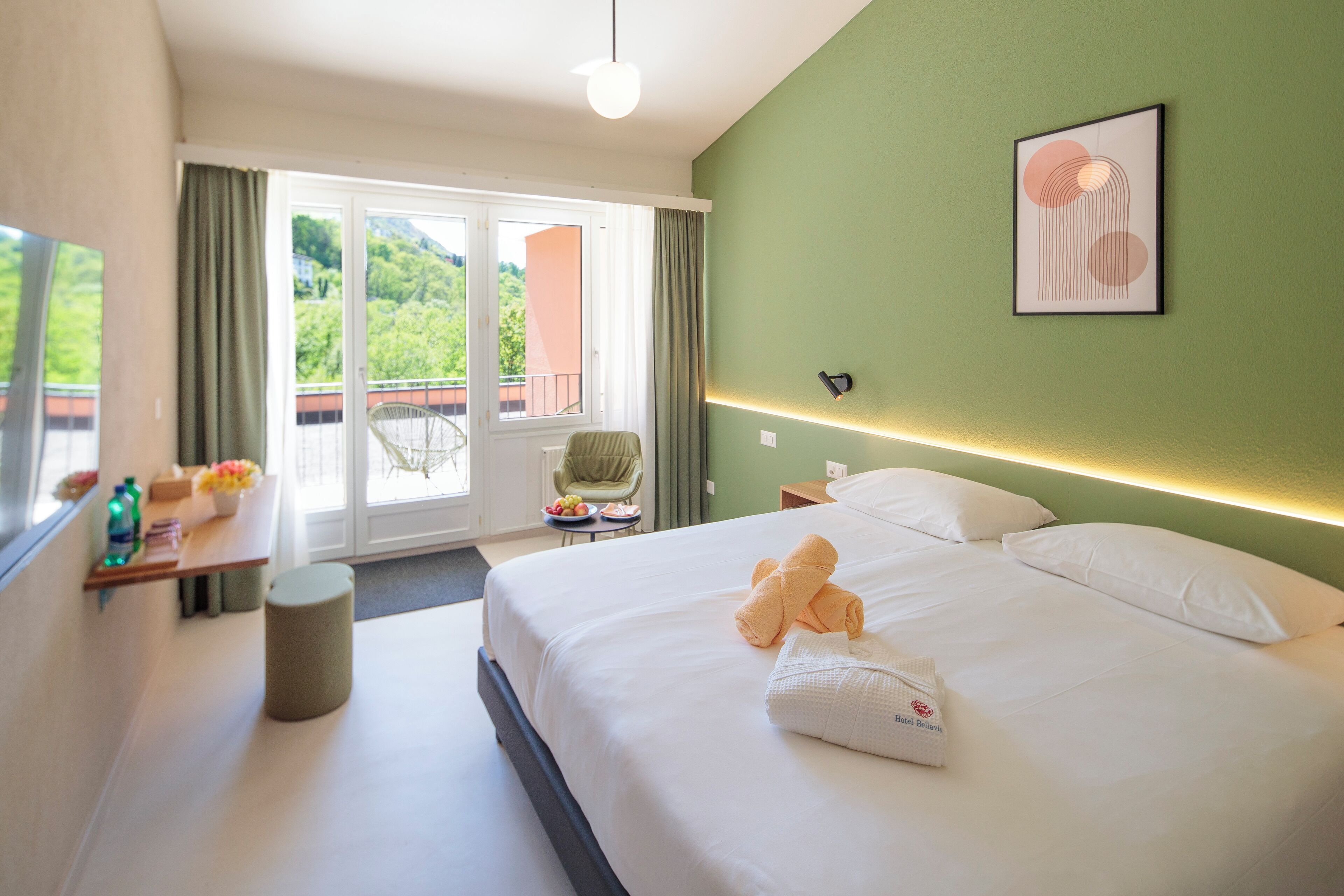 A hotel room with a bed, a chair, a small table with a bowl of fruit, and a view of greenery outside.