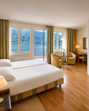 Une chambre d’hôtel avec un grand lit, deux fauteuils, un bureau et une vue sur un lac et des montagnes.