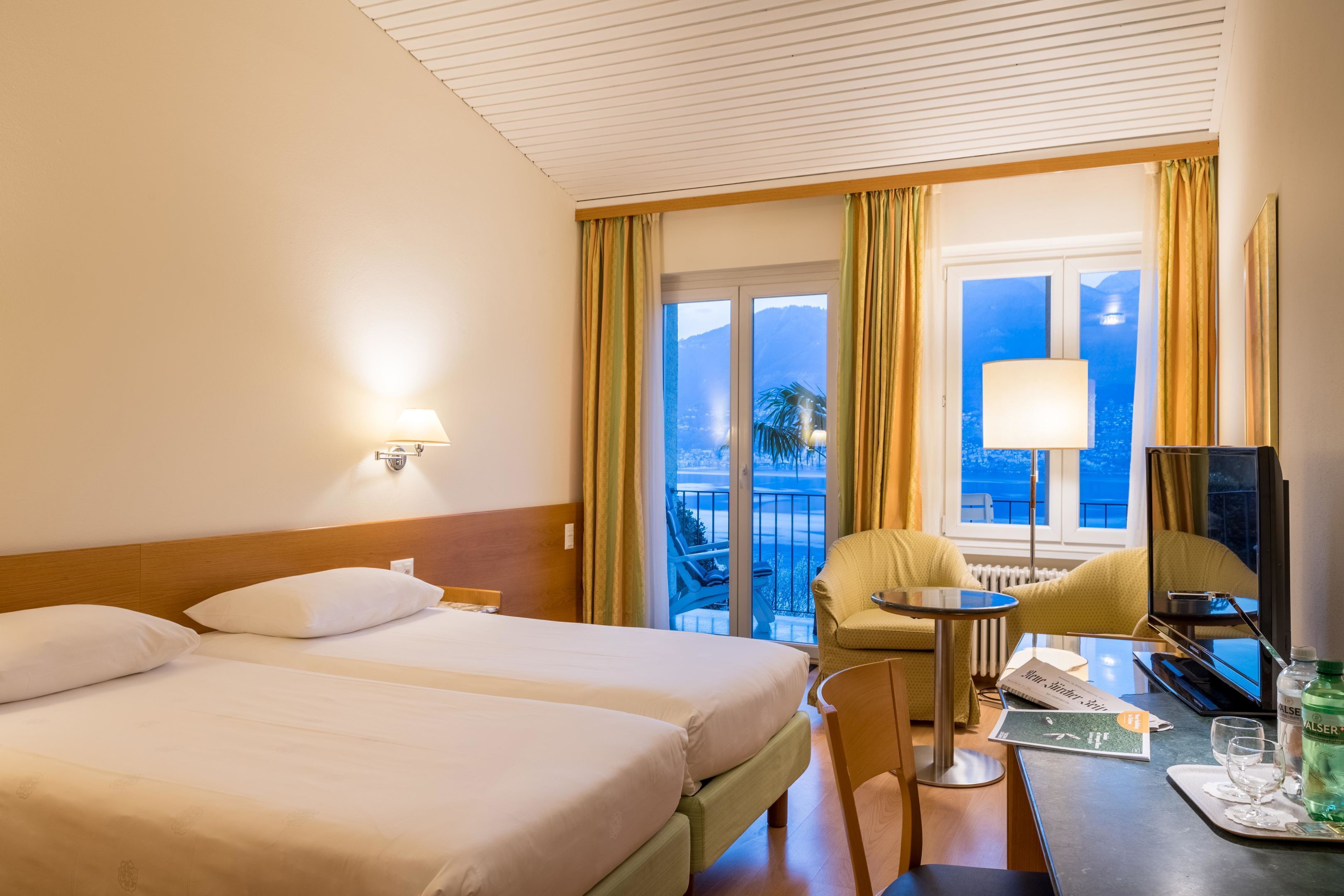 A hotel room with two beds, a desk with a lamp, a chair, a TV, and a view of the outdoors.