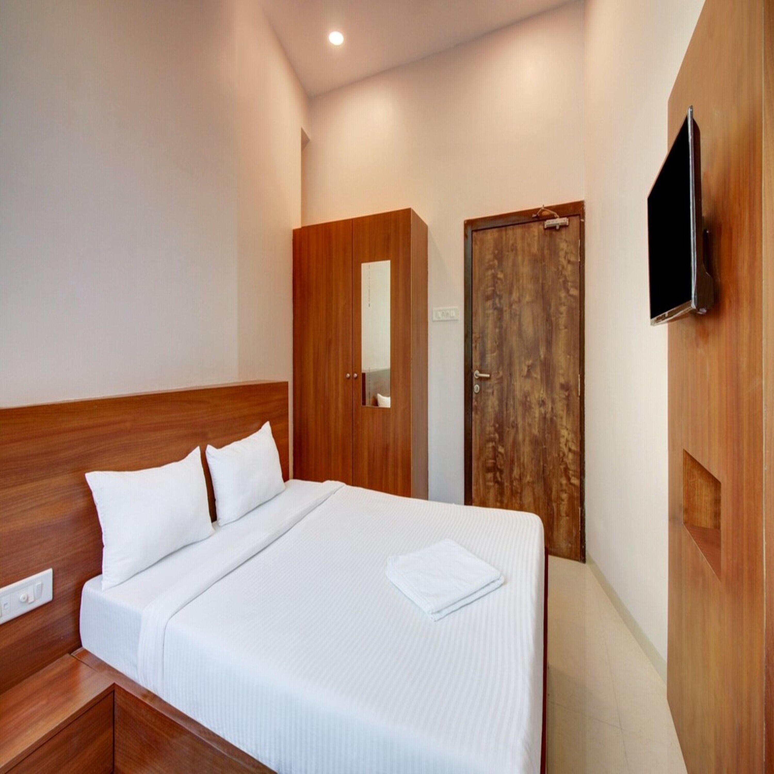 A neatly made bed with white linens and pillows, a wooden headboard, a wooden wardrobe with a mirror, and a flat-screen TV mounted on the wall.