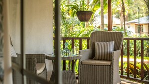 A wicker chair with a cushion and a glass table with a bottle and glass on a balcony with a view of greenery. - Karen Blixen Coffee Garden & Cottages (Nairobi)