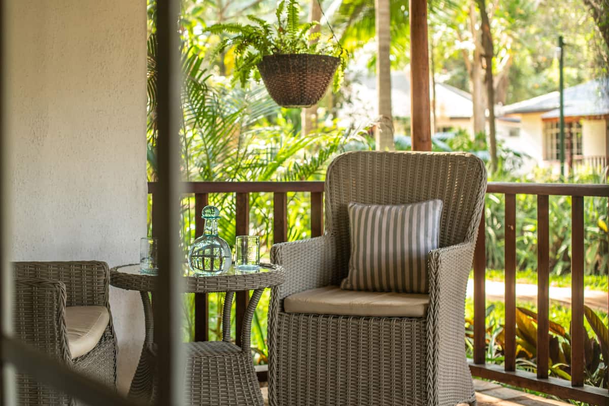 A wicker chair with a cushion and a glass table with a bottle and glass on a balcony with a view of greenery.