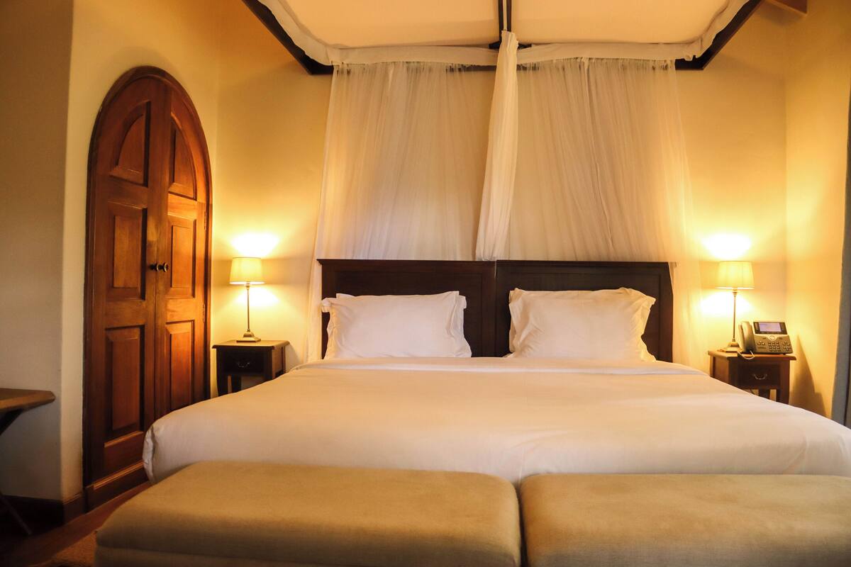 A neatly made bed with white linens, two bedside lamps, a wooden door, and a canopy overhead.