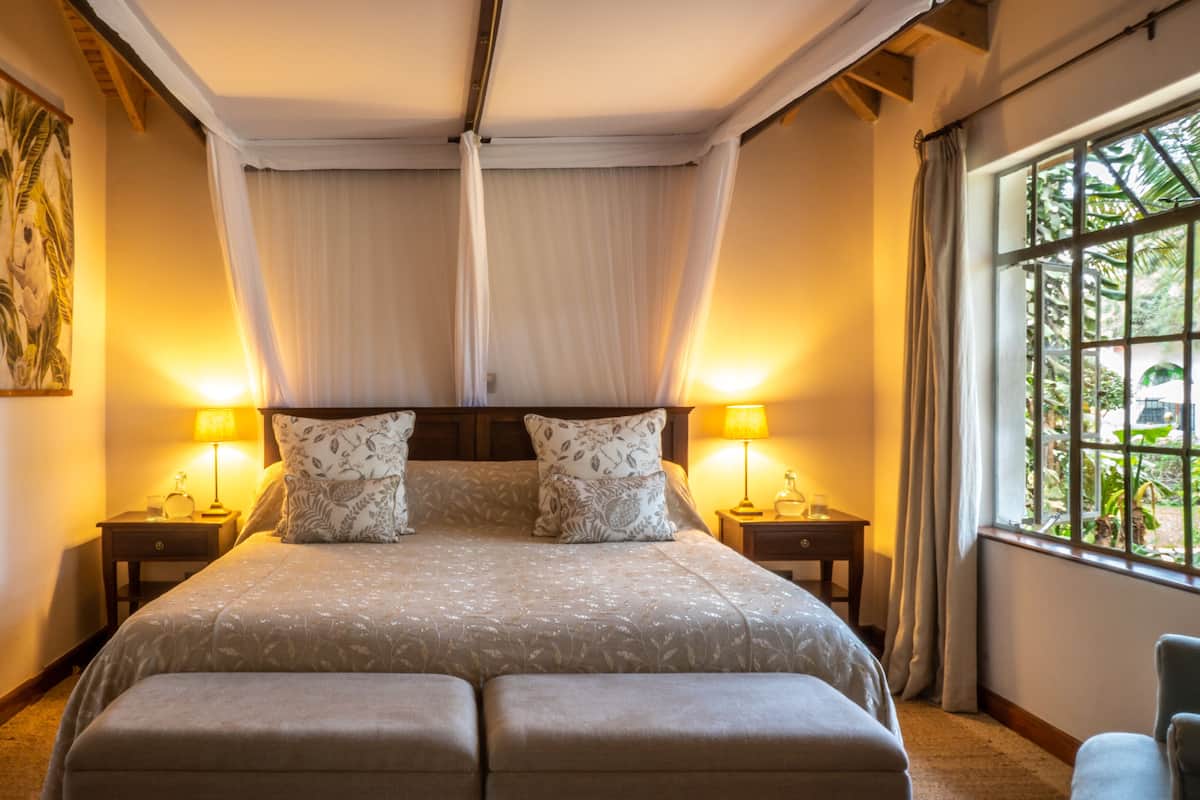 A bedroom with a large bed, two bedside tables with lamps, a canopy bed, a window with a view of greenery, and a painting on the wall.