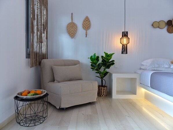 A modern bedroom with a sofa, a bedside table with a lamp, a potted plant, and a fruit basket.