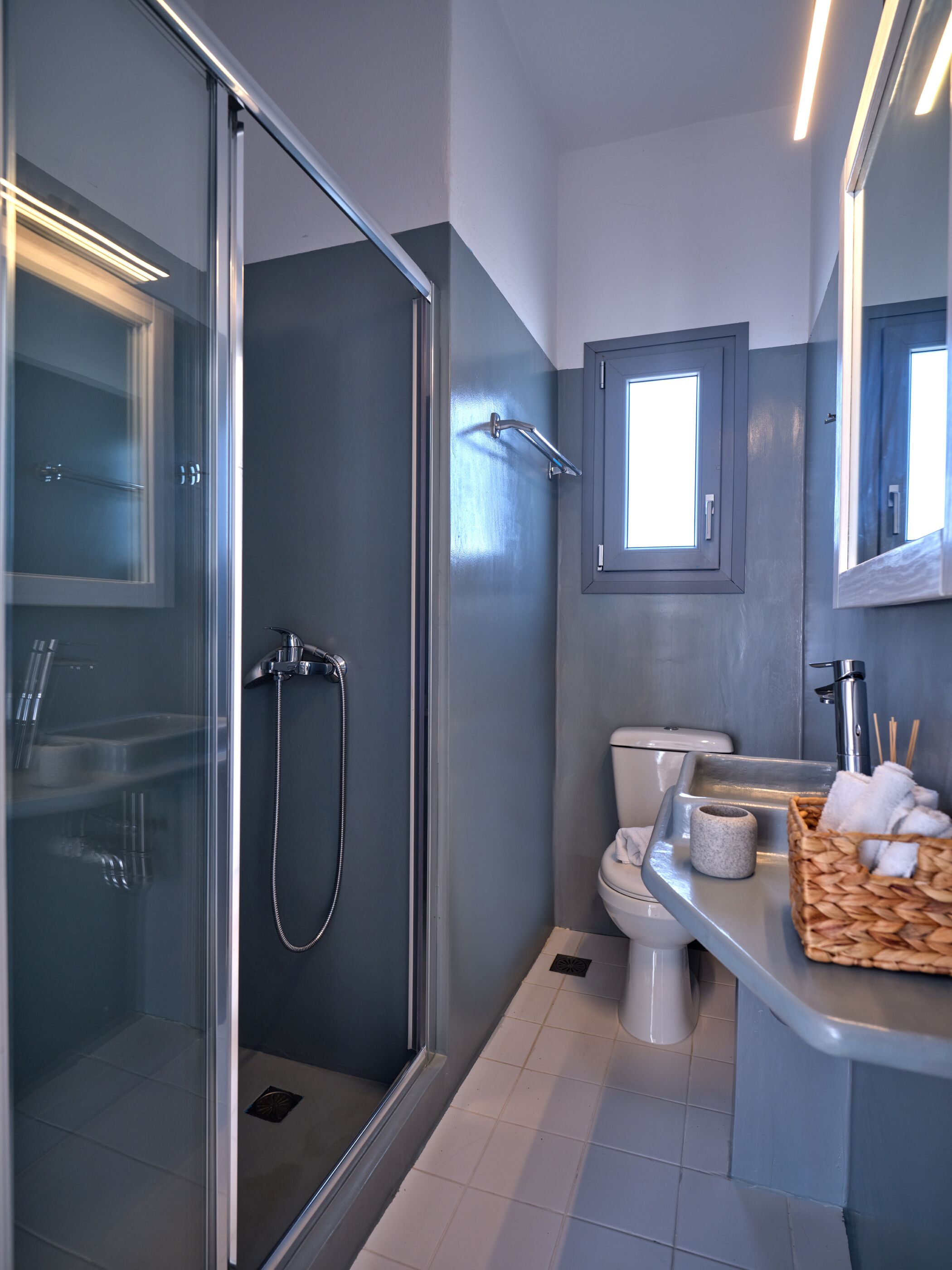 Un baño moderno con ducha envuelta en vidrio, inodoro y lavamanos con un cesto de artículos de aseo.