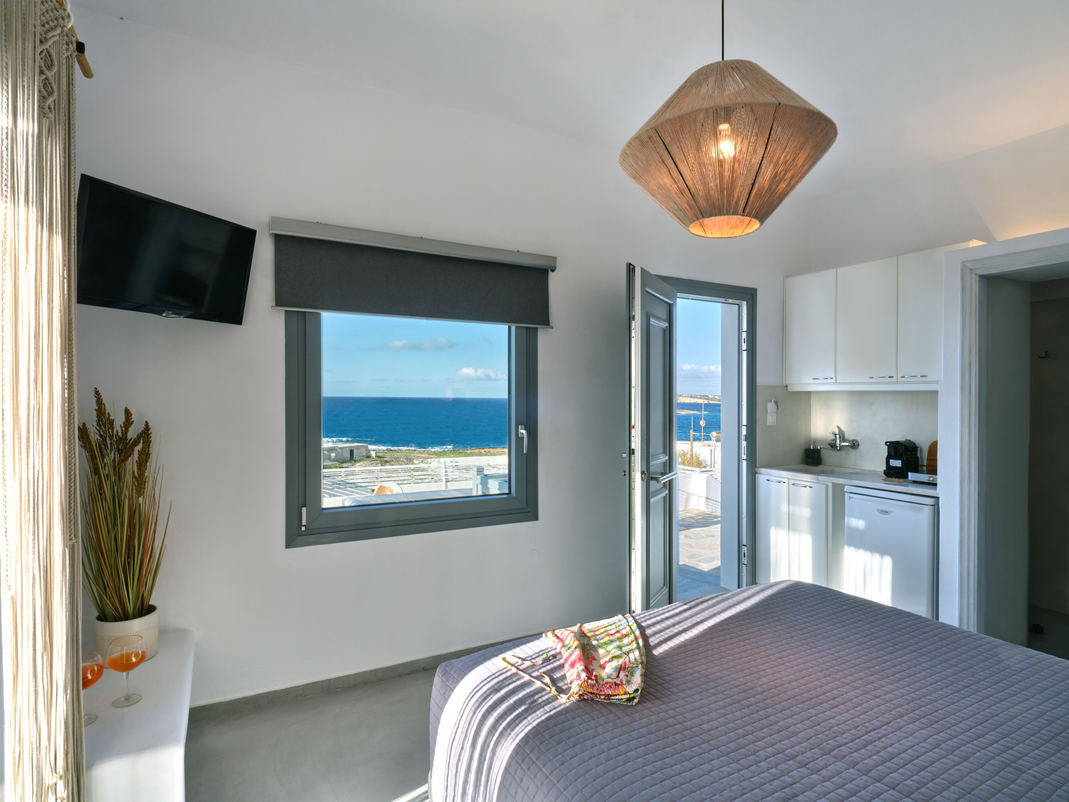 A bedroom with a large bed, a TV mounted on the wall, a window with a view of the sea, and a kitchen area visible through an open door.
