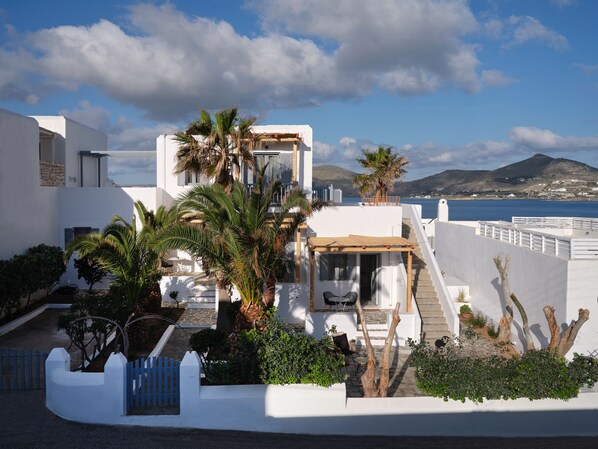 A modern white building with a patio area, palm trees, and a view of the sea and hills. - Ariti Seaside Residence (Paros)