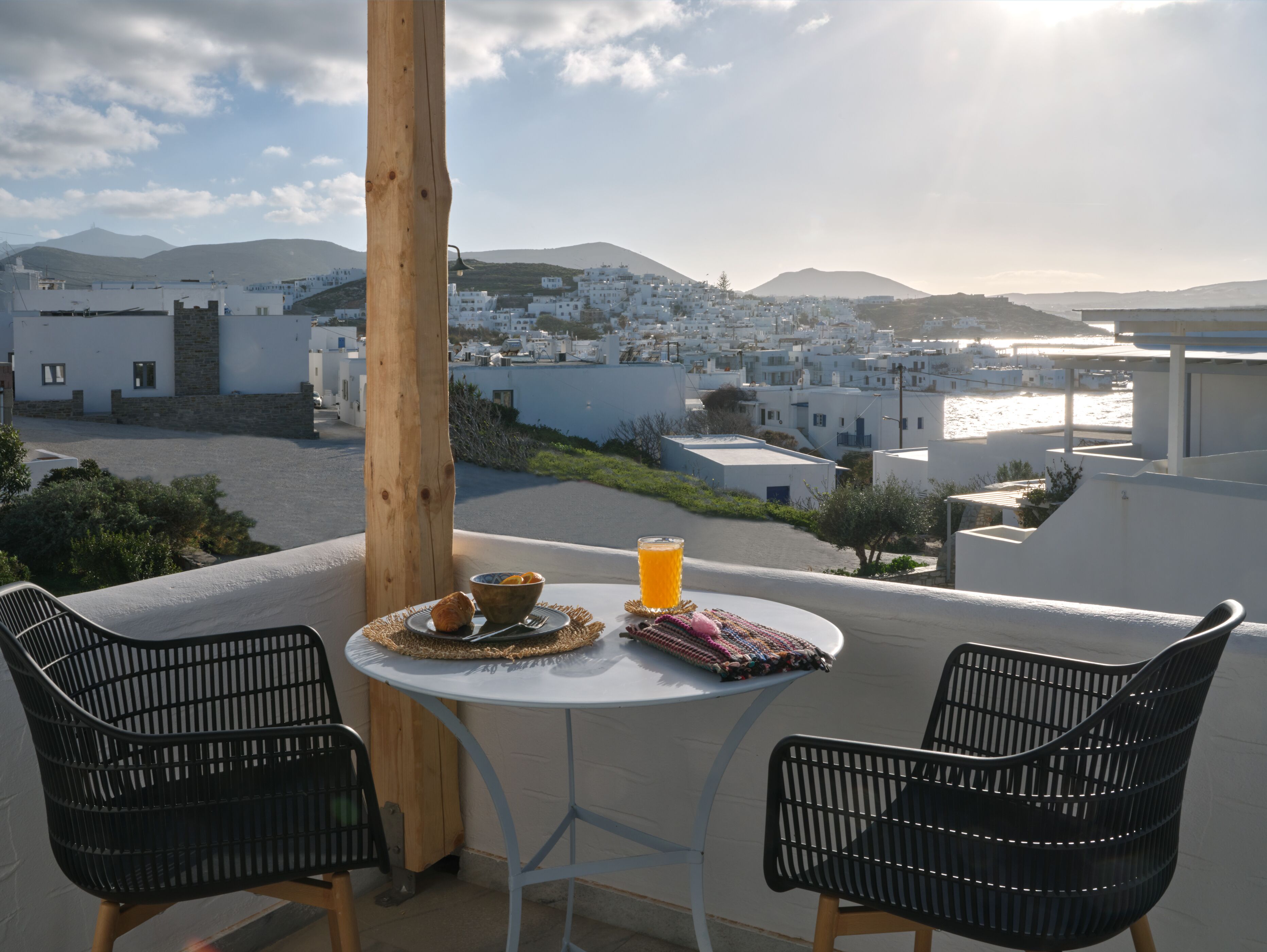 A balcony with two black chairs, a small round table with a bowl of fruit, a glass of orange juice, and a candle. The view includes white buildings and a mountain in the distance.