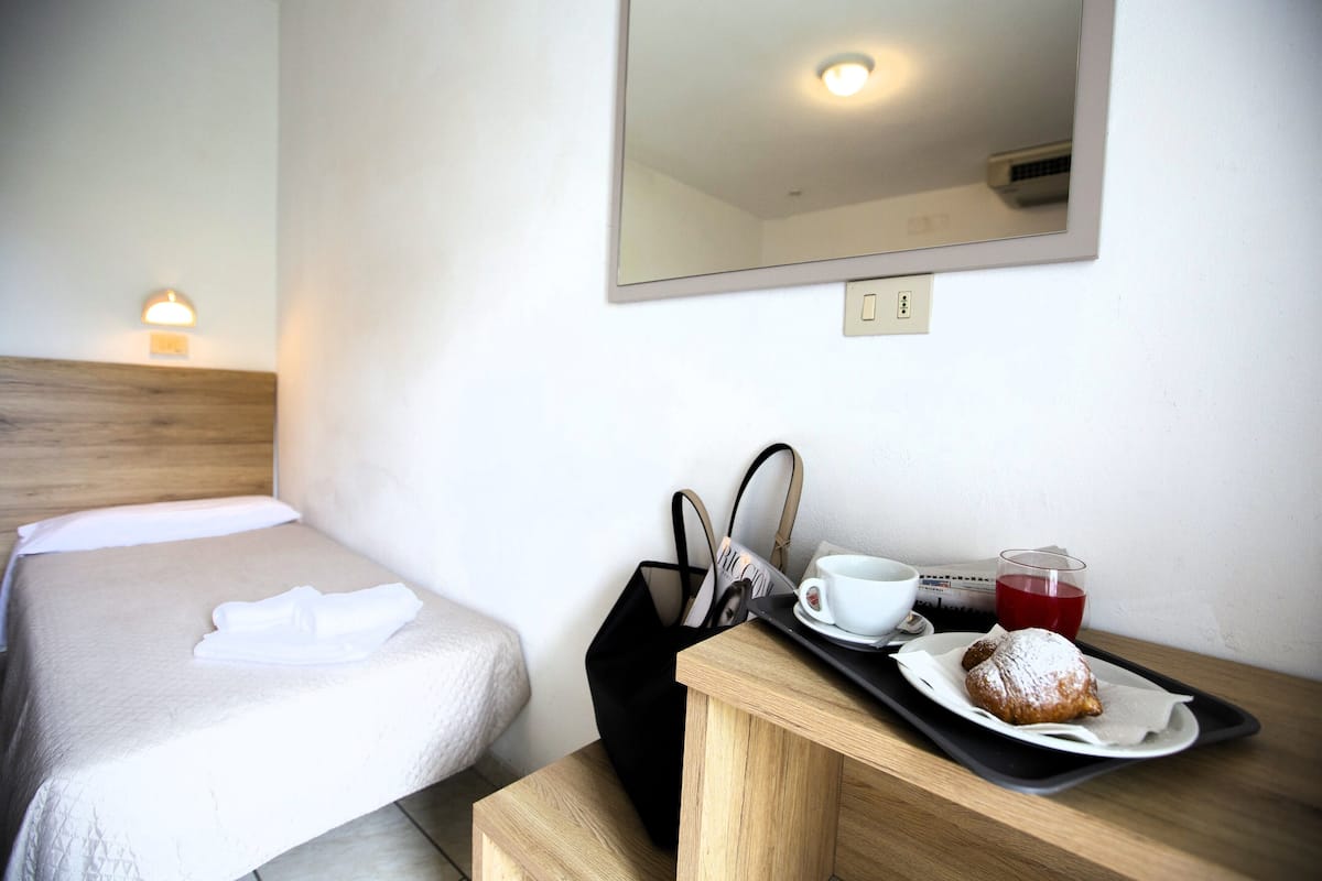 A hotel room with a bed, a bedside table with a tray of food, a cup, and a bag.