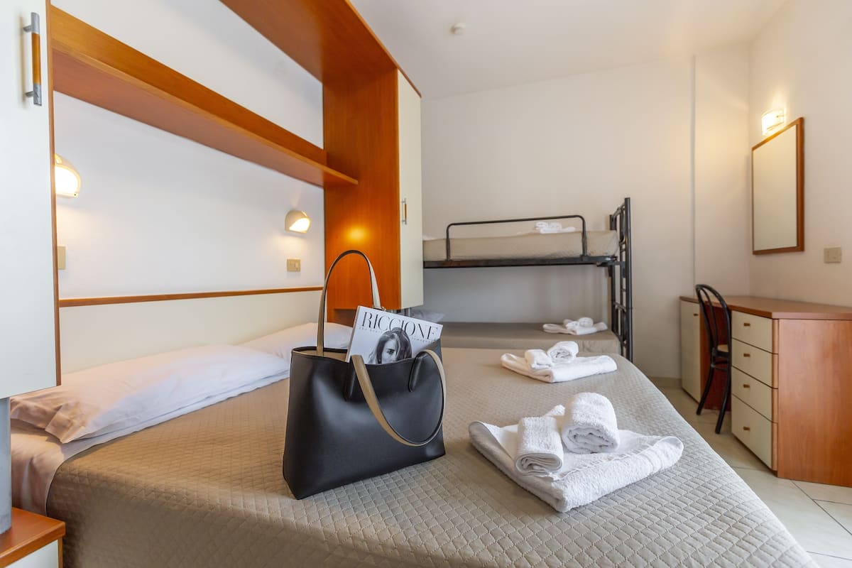 A hotel room with a bunk bed, a bedside table, a wooden wardrobe, and a black handbag with a magazine on it.